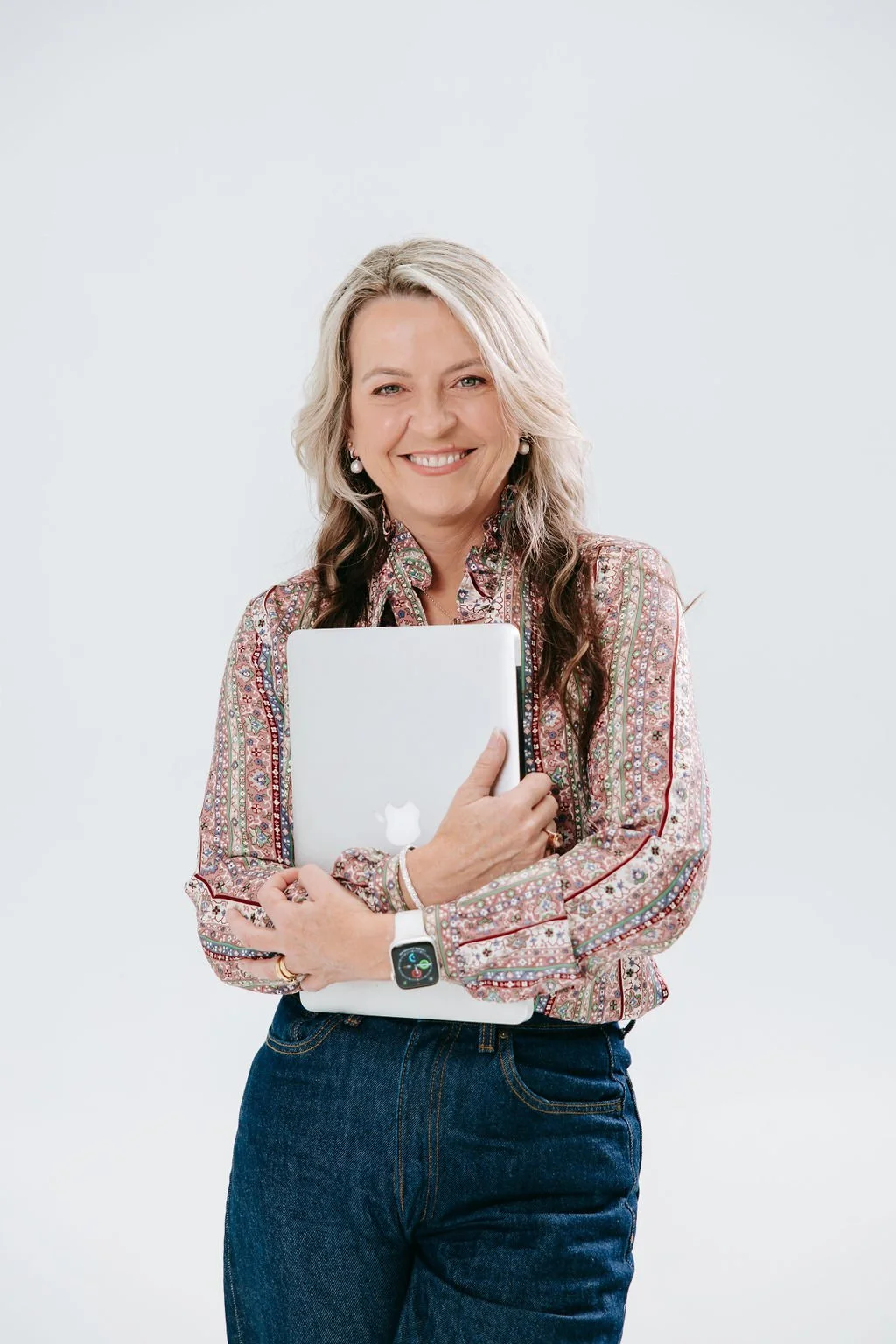 A Picture featuring Psychologist Kerstin Anderson Ridge - A smiling woman with blonde hair, wearing a patterned blouse and jeans, holding a closed MacBook and wearing an Apple Watch against a plain white background.