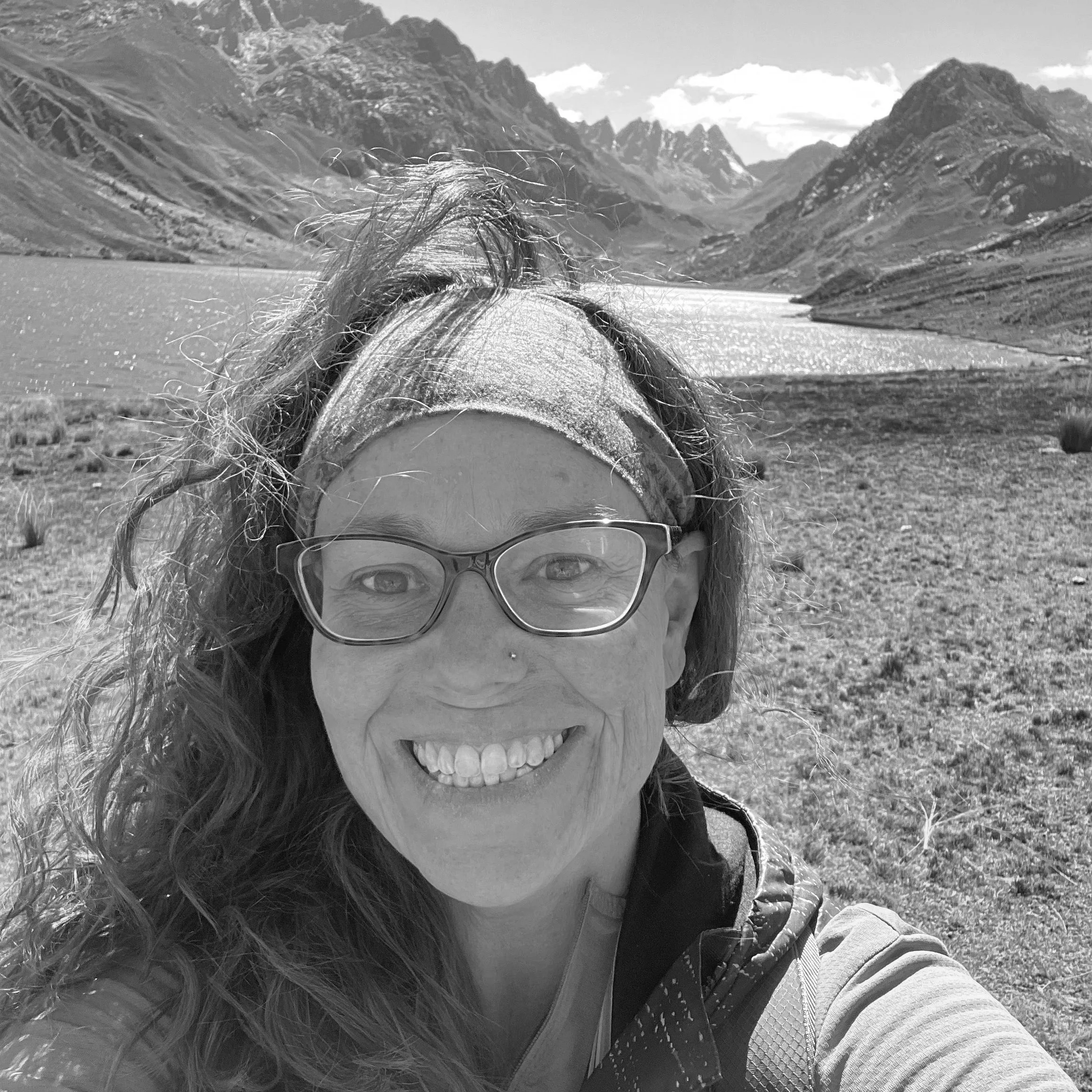 A person wearing sunglasses and a jacket with a hood, smiling, in front of mountain and lake scenery.