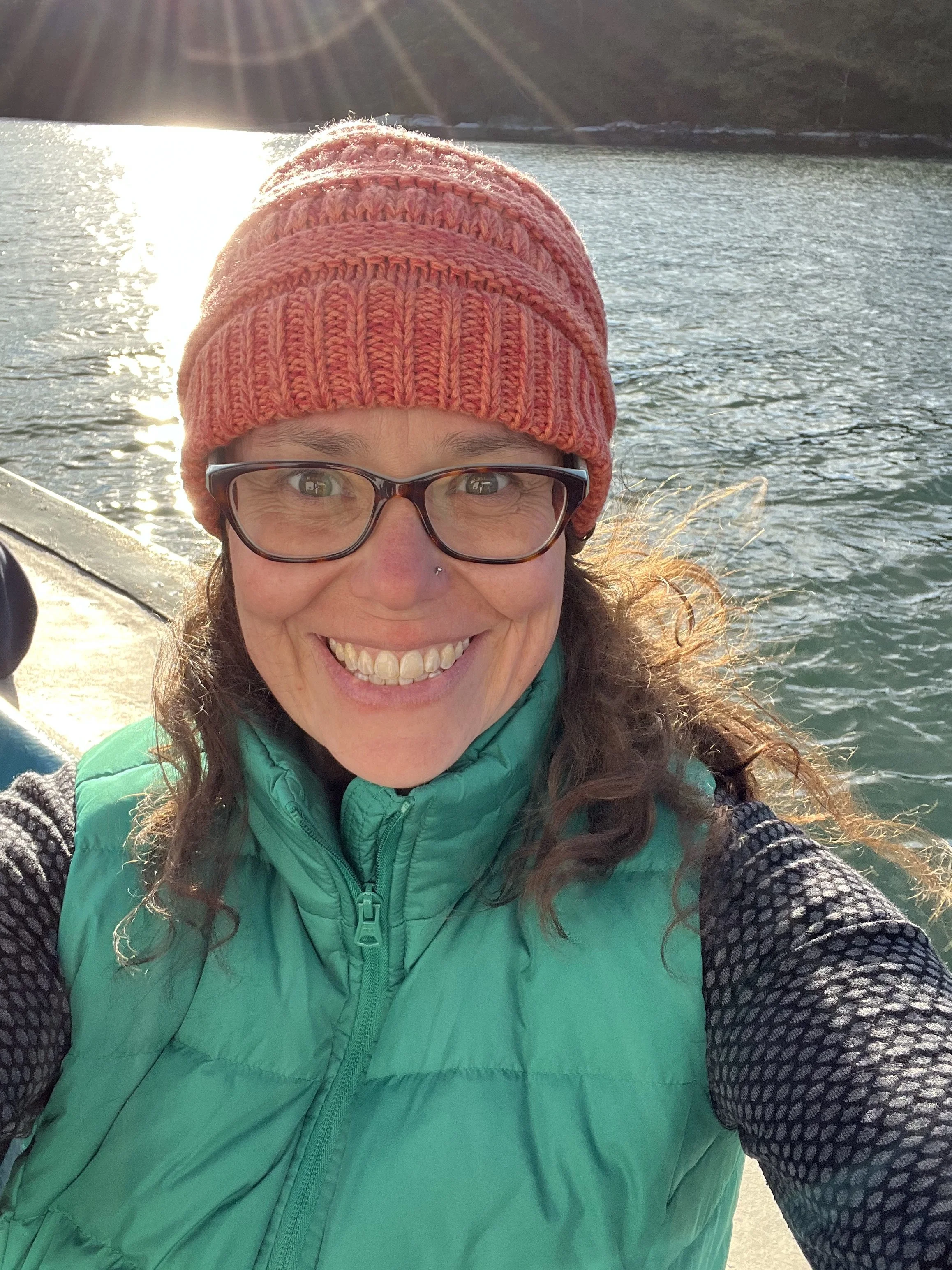 A woman smiling outdoors near a lake, wearing glasses, a pink knit beanie, a green puffer vest, and black patterned long sleeves, with sunlight reflecting on the water in the background.
