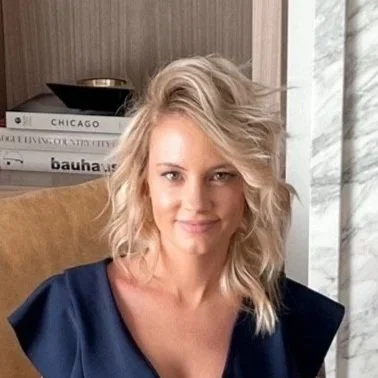 A woman with shoulder-length blonde hair, wearing a navy blue top, sitting indoors in front of a stack of books and a curtain.