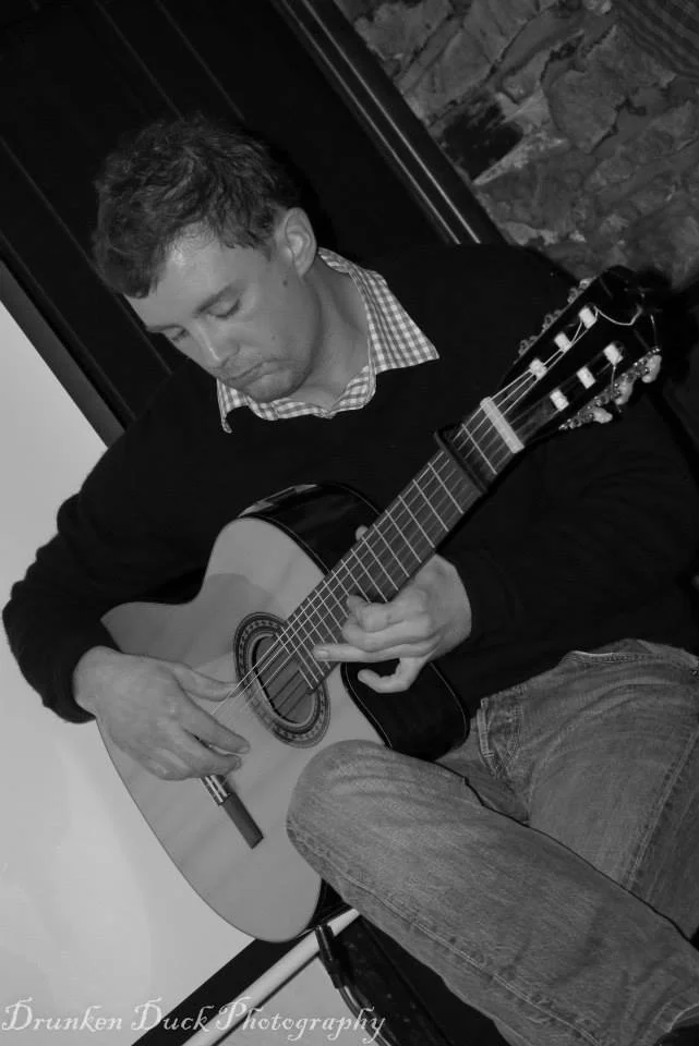 A man playing an acoustic guitar, sitting indoors with a stone wall in the background, black and white photo.