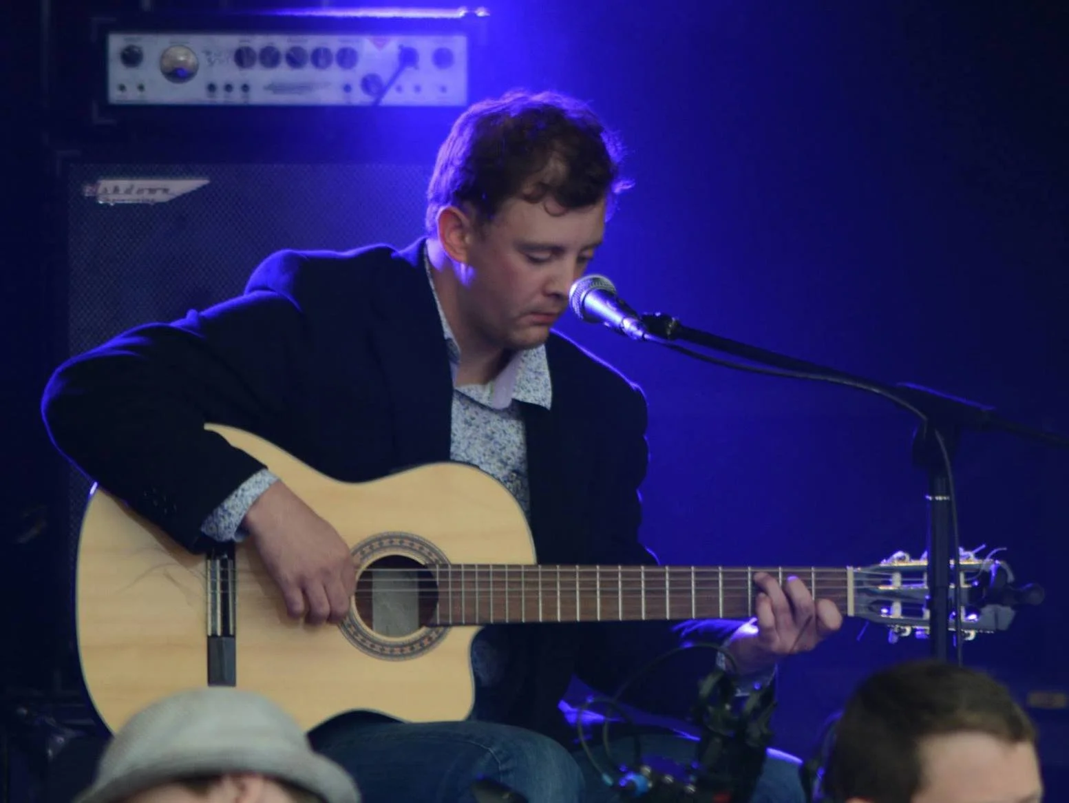 A man playing an acoustic guitar on stage, singing into a microphone with purple and blue stage lighting.