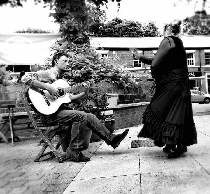 A young man playing guitar while sitting on a wooden chair and a woman dancing outdoors on a sidewalk during the daytime.