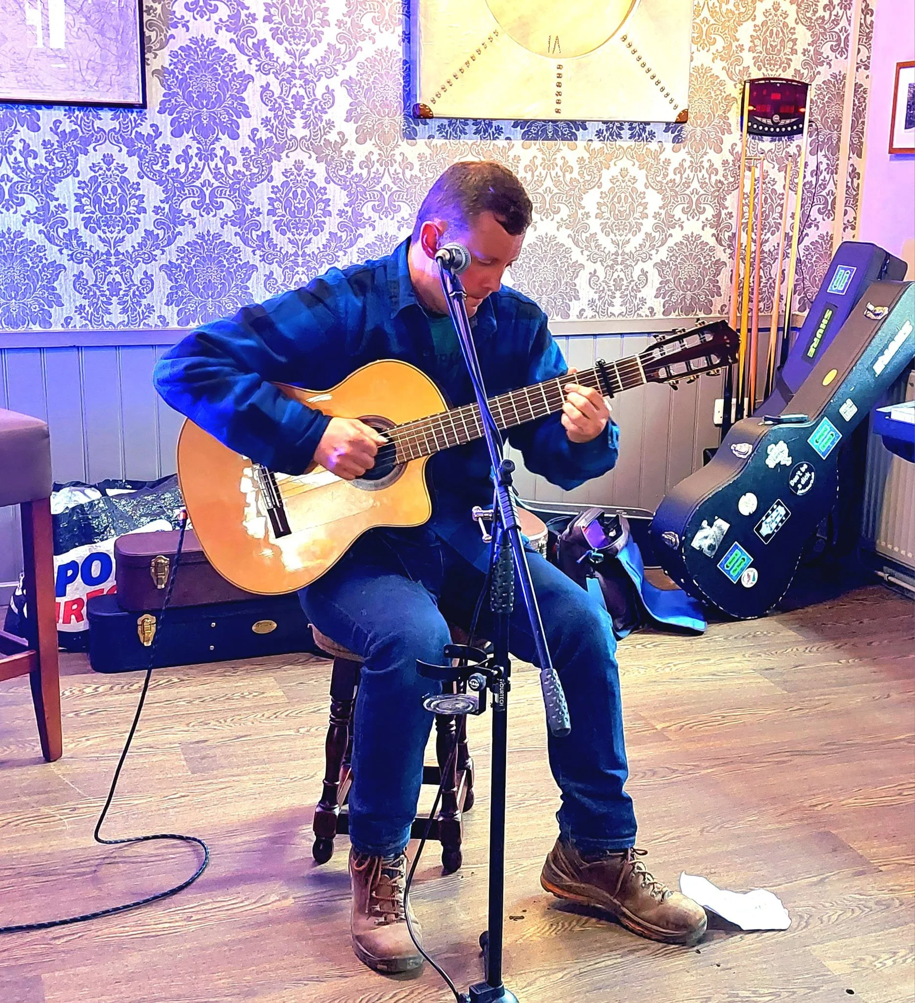 A young man sitting on a wooden stool playing an acoustic guitar, with a microphone in front of him. He is wearing brown boots, blue jeans, a blue plaid shirt, and a teal t-shirt. The room has patterned wallpaper with a gold and purple design, framed