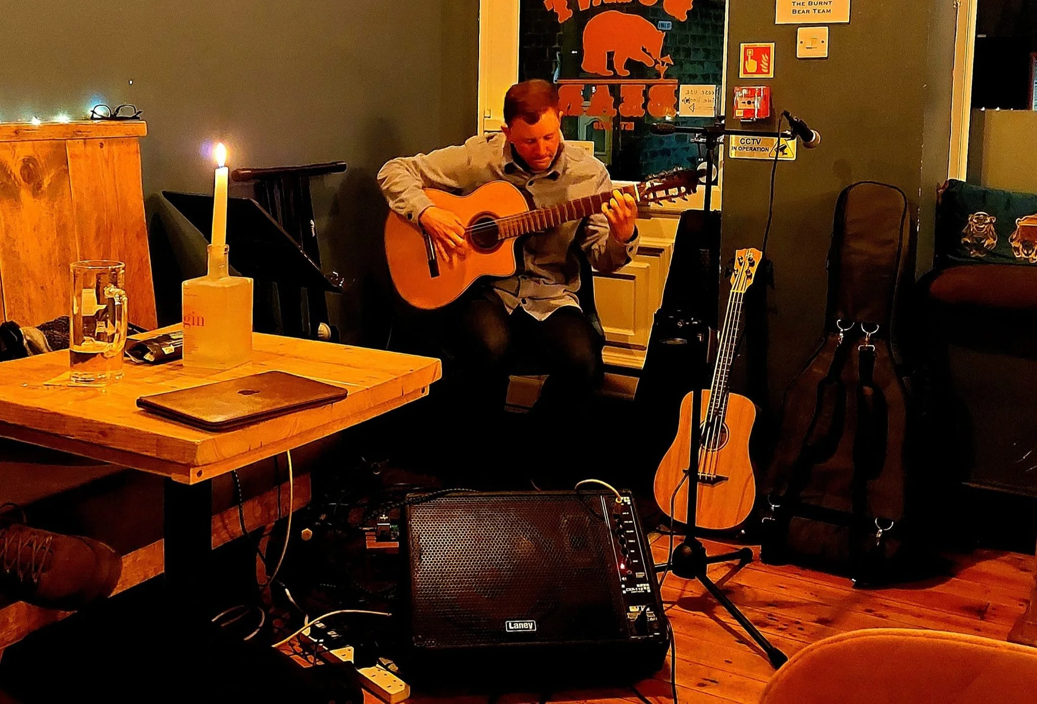 A man playing an acoustic guitar indoors, surrounded by musical equipment and chairs, with a poster of a bear on the wall behind him. The scene is warmly lit, suggesting an intimate performance or practice session.
