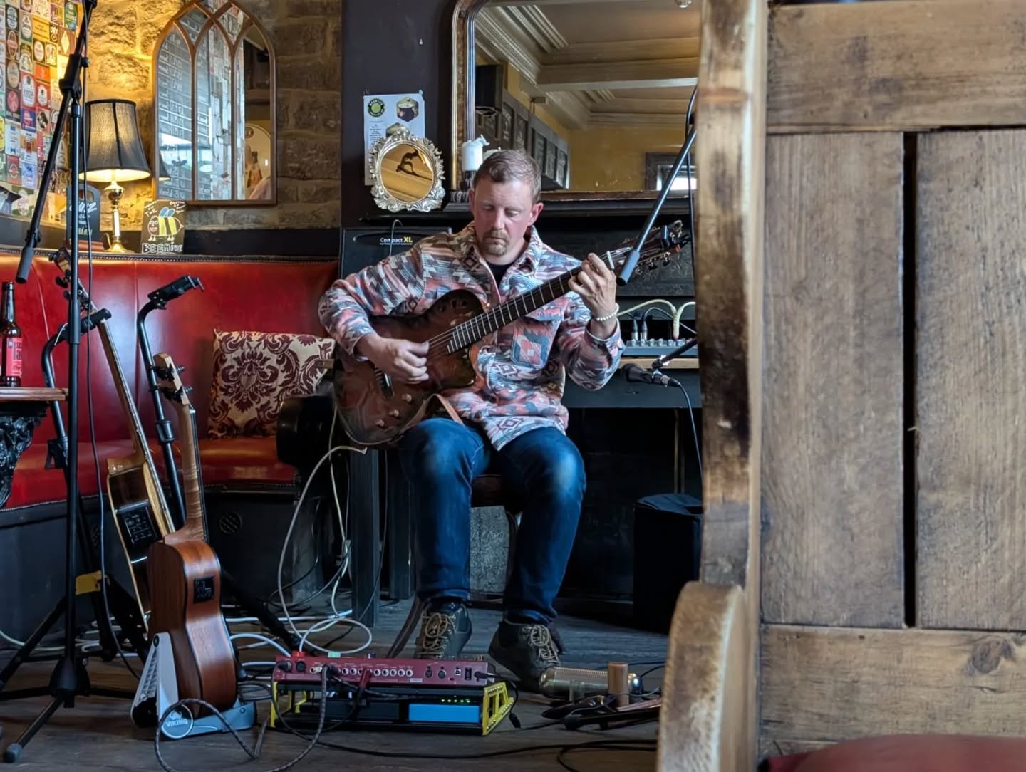 A man playing an acoustic guitar in a cozy, rustic pub or restaurant setting, surrounded by musical equipment and instruments.