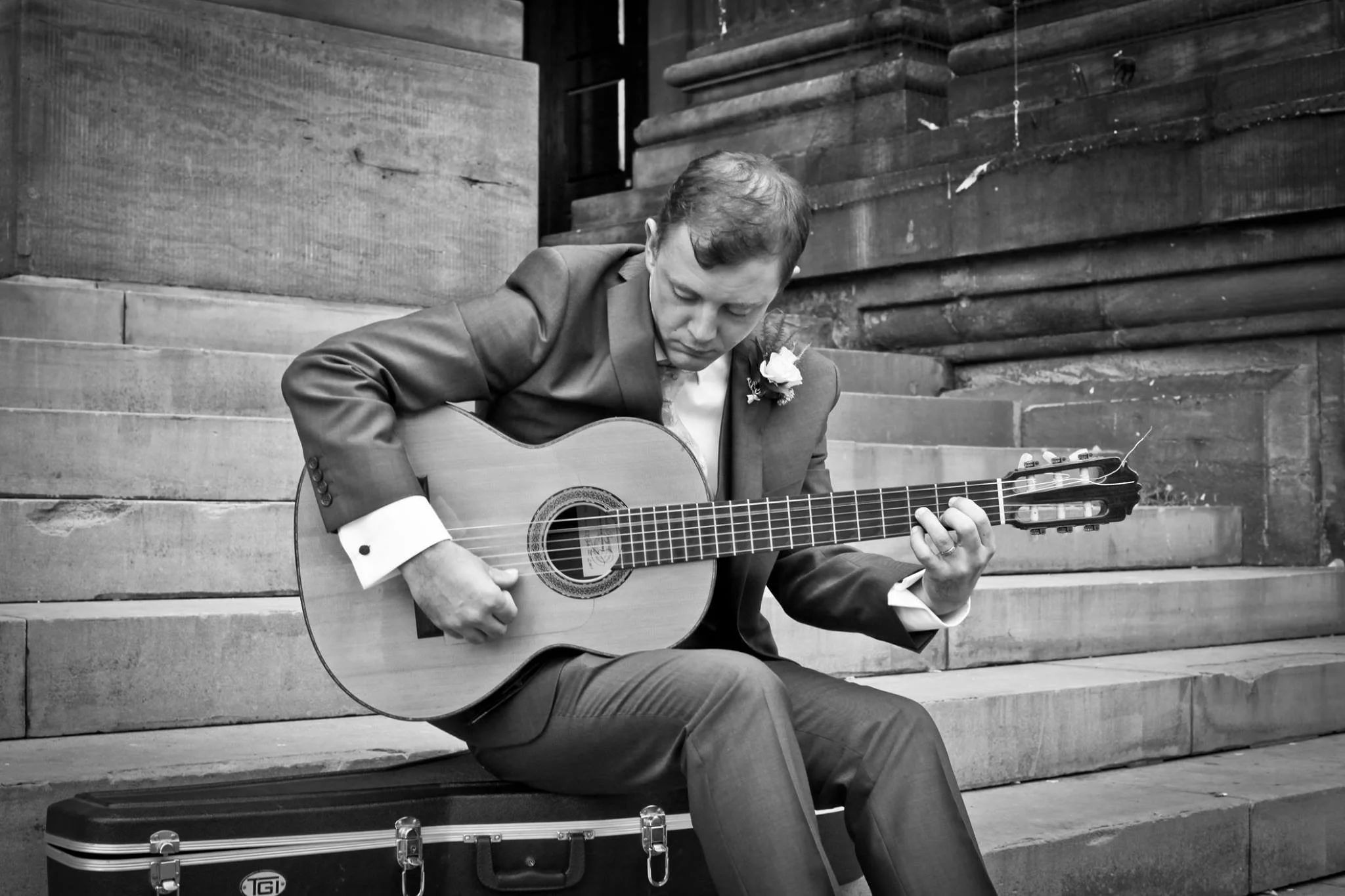 Black and white photo of a man in a suit playing an acoustic guitar, seated on steps outside. He has a flower on his lapel and is looking down at the guitar. There is a guitar case on the steps beside him.