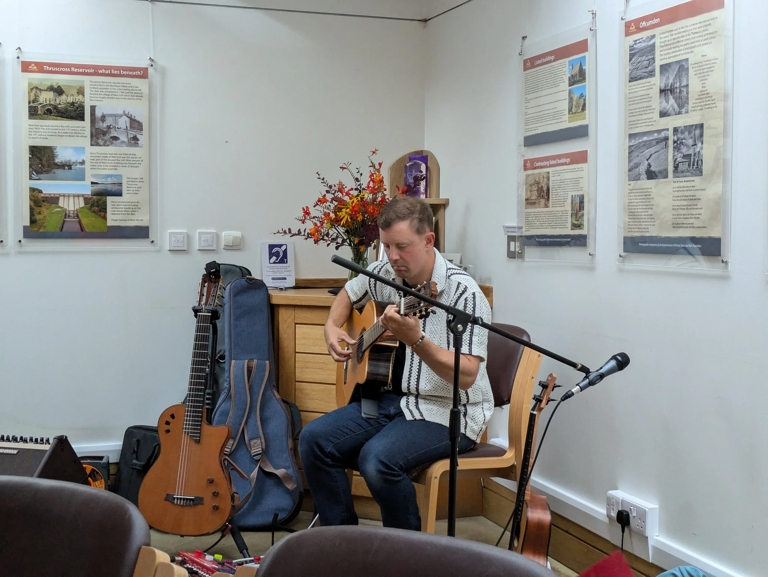 Man playing acoustic guitar, sitting on a wooden chair in a room with informational posters on the wall, a flowering plant, and musical equipment including another guitar, a microphone, and a keyboard.