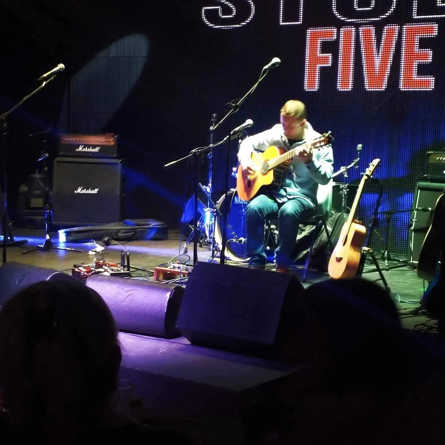 A musician sitting on a stage playing an acoustic guitar, surrounded by music equipment, microphones, and amplifiers. A sign in the background reads 'FIVE'.