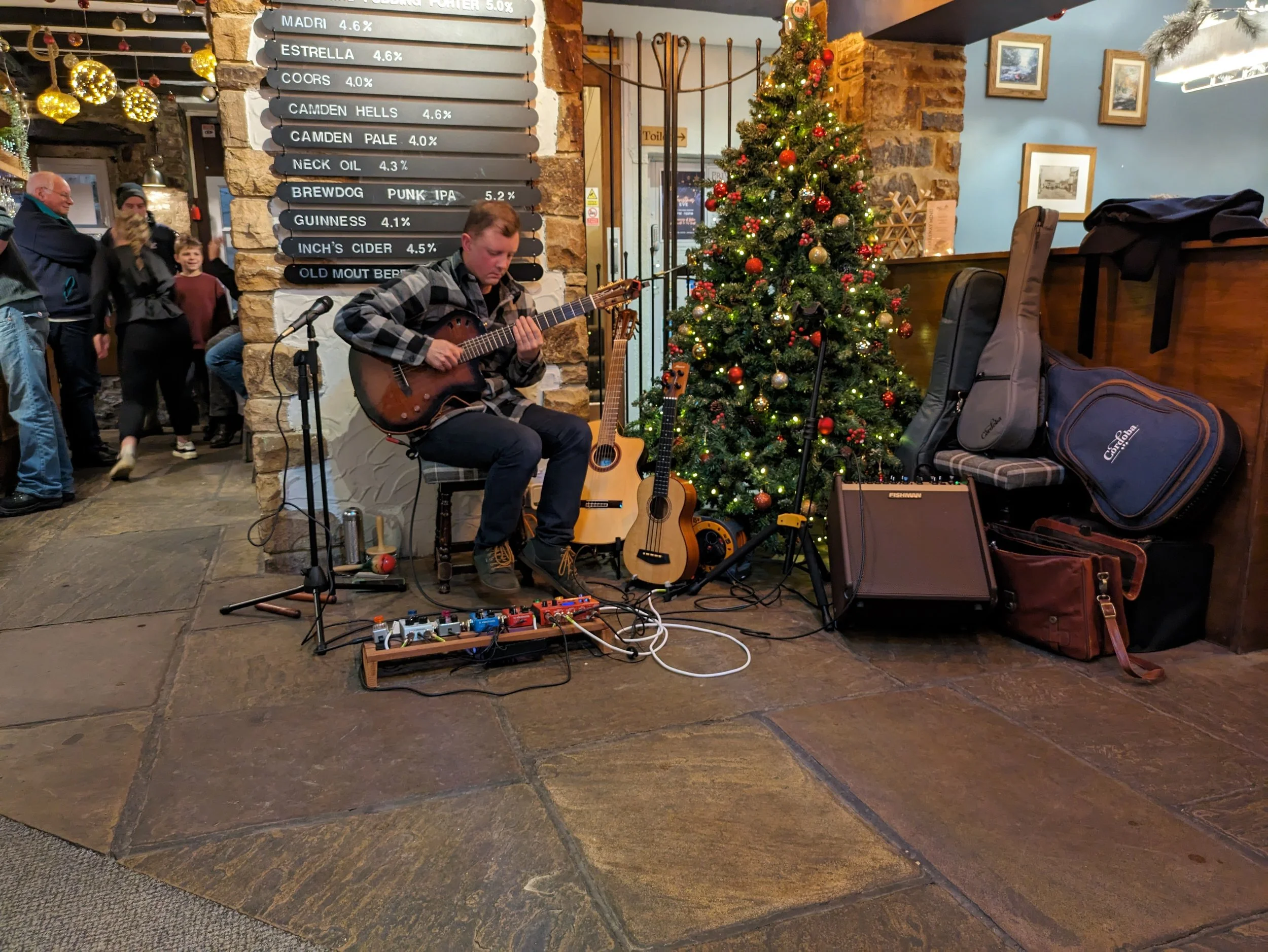 A person playing an acoustic guitar in front of a decorated Christmas tree in a cozy indoor setting with a group of people watching and smiling.