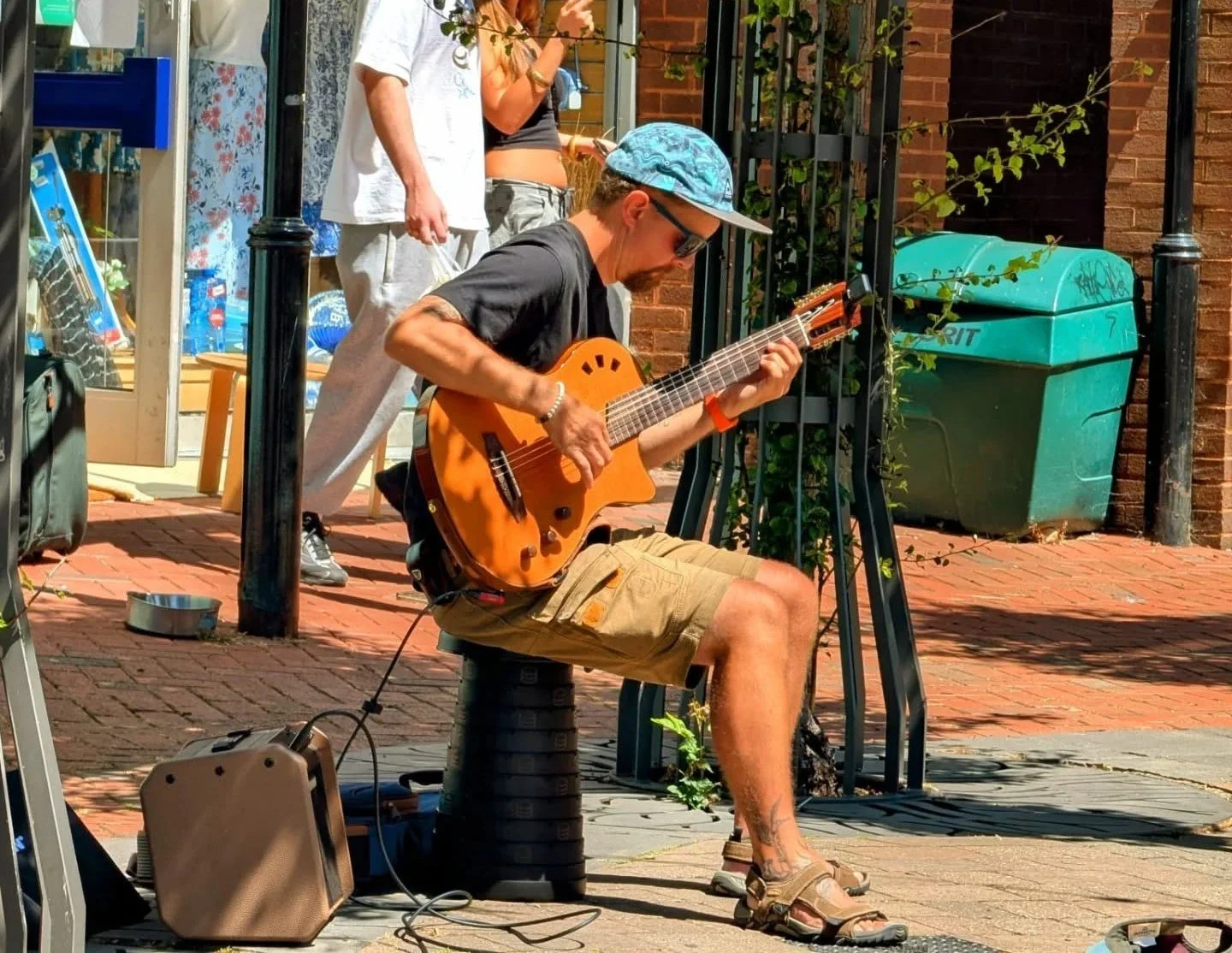 A man sitting on a stool playing an acoustic guitar on a sidewalk, with a woman standing nearby and a person in the background. The man is wearing sunglasses, a blue cap, a black t-shirt, tan shorts, and sandals. There is a small amplifier on the gro