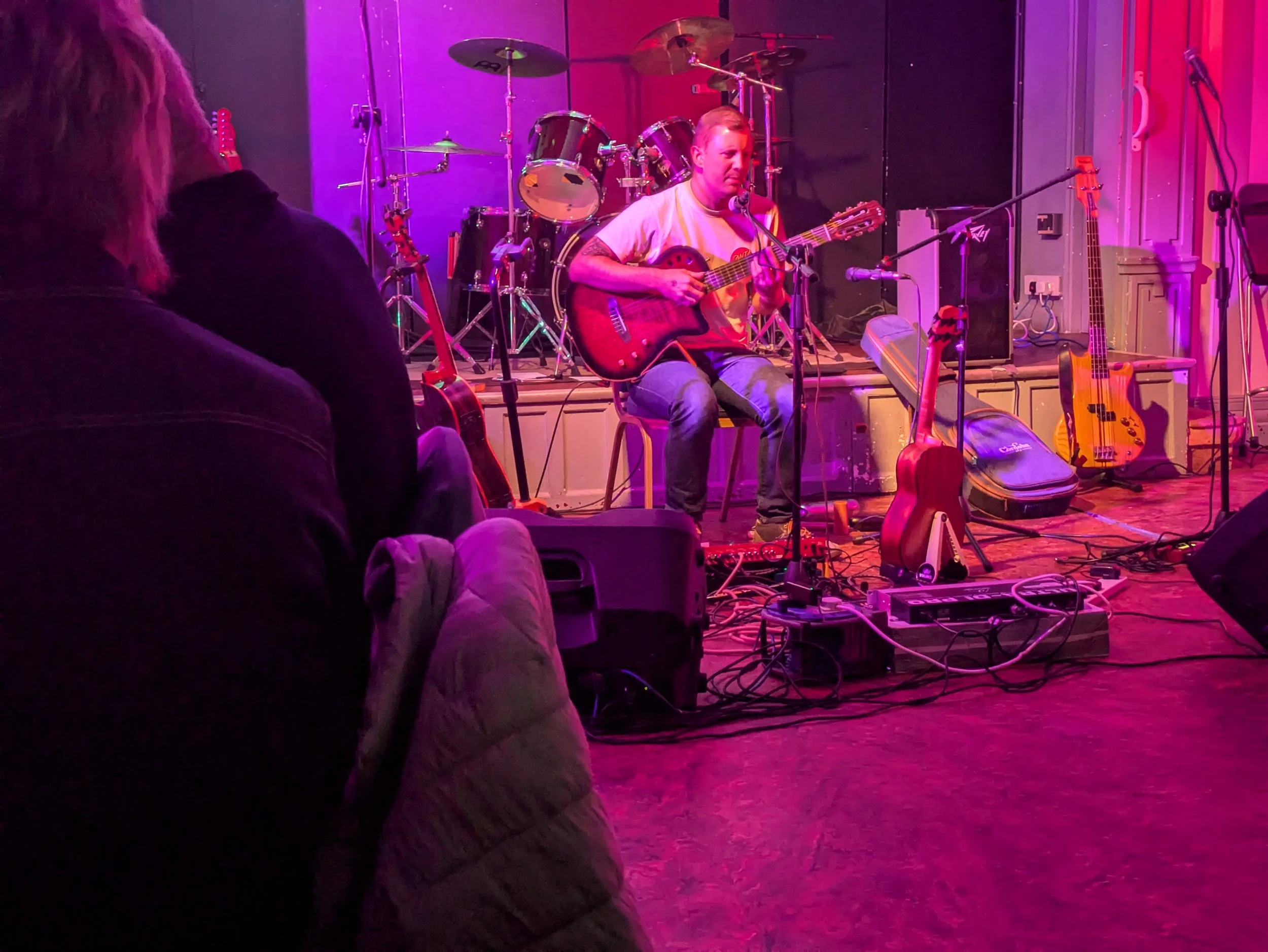 A musician playing an acoustic guitar on a stage with drums and other instruments, illuminated by pink and purple stage lighting, with an audience member visible in the foreground.
