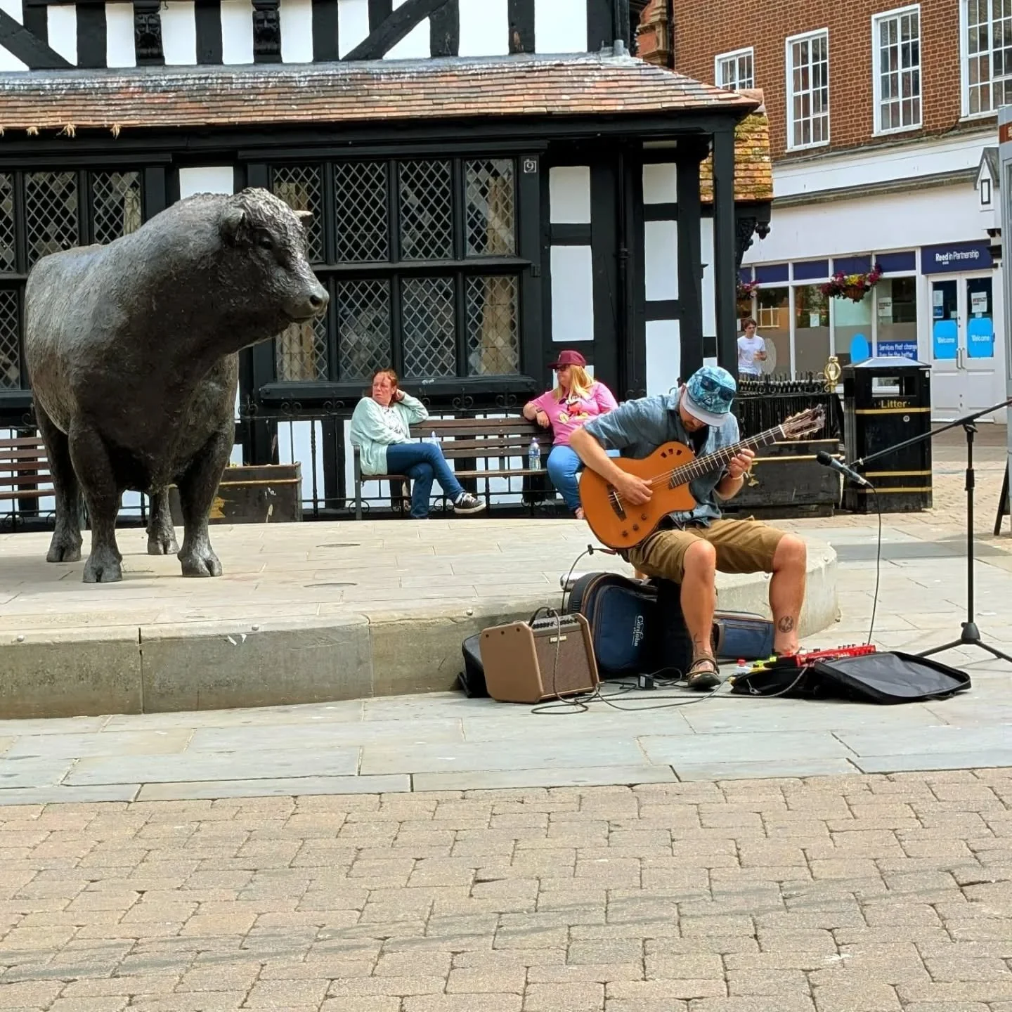 A street musician playing guitar and singing into a microphone on a sidewalk with a bronze boar statue nearby. Two women are sitting on benches in the background, one wearing a white jacket and the other in a pink shirt. A person in a white shirt is 