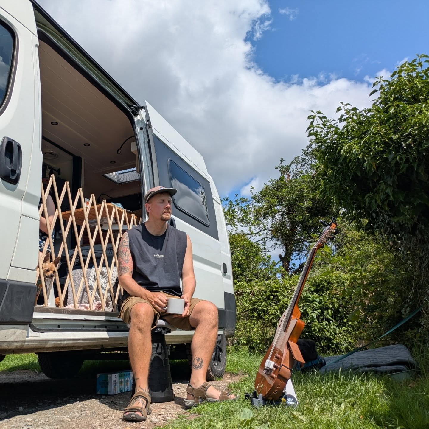 A man sitting outside a camper van, holding a mug, with a guitar in a stand nearby, a woman with a dog inside the van, surrounded by greenery and blue sky.