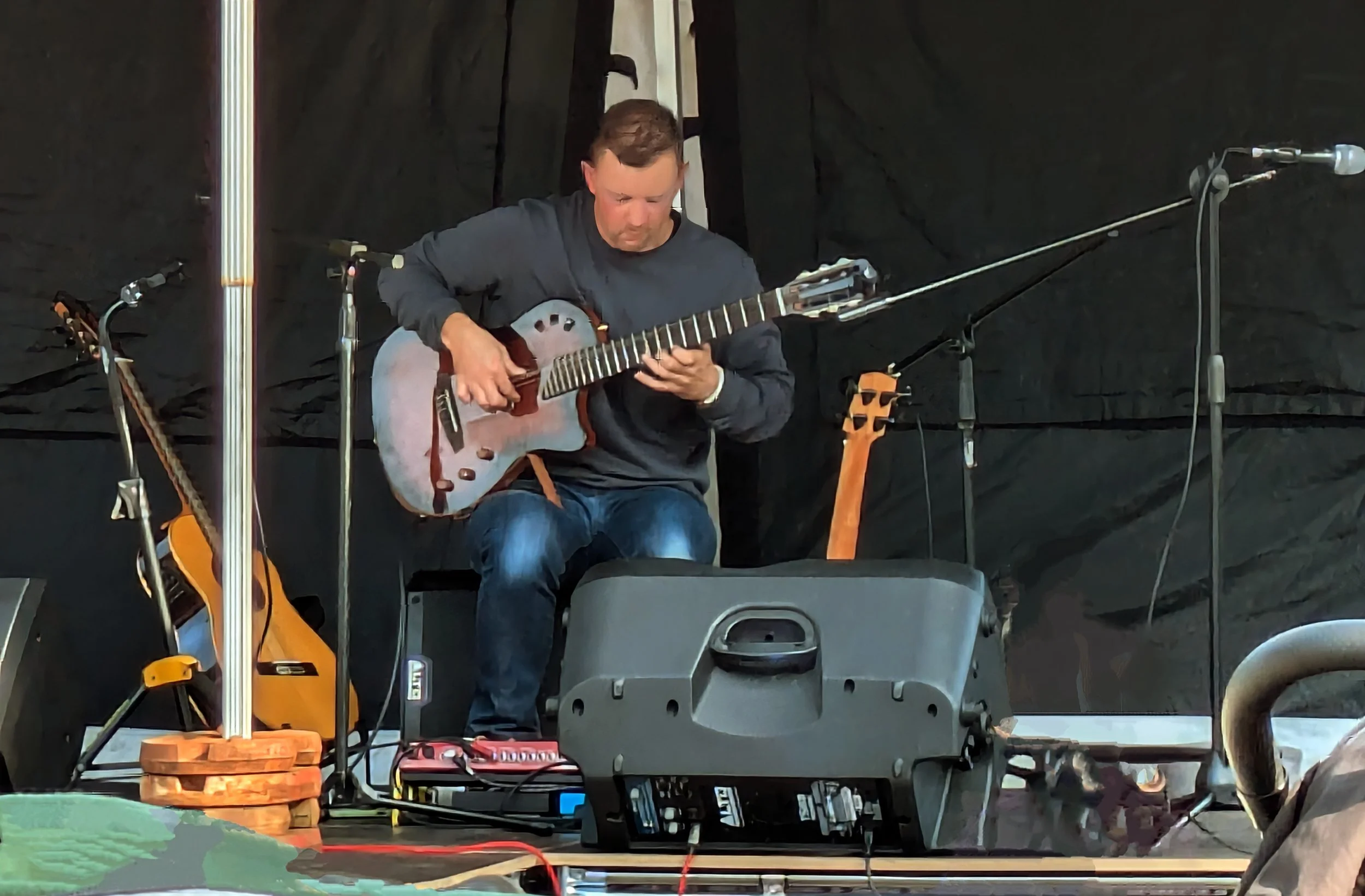 A man sitting outdoors on a stage playing an acoustic guitar. He is surrounded by microphone stands, other guitars, and audio equipment with a black backdrop.