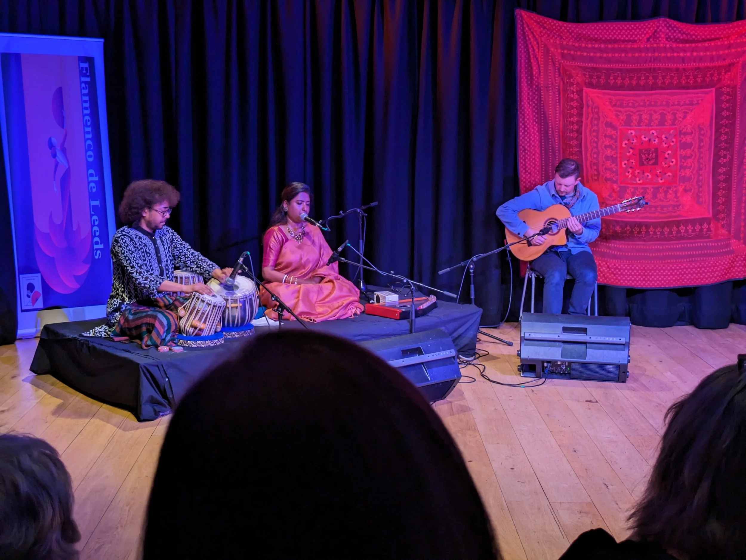 A live musical performance on stage with three performers: a male with glasses playing drums, a woman singing into a microphone, and a man playing guitar. The stage has a navy curtain backdrop and a red patterned tapestry on the right. A banner with 