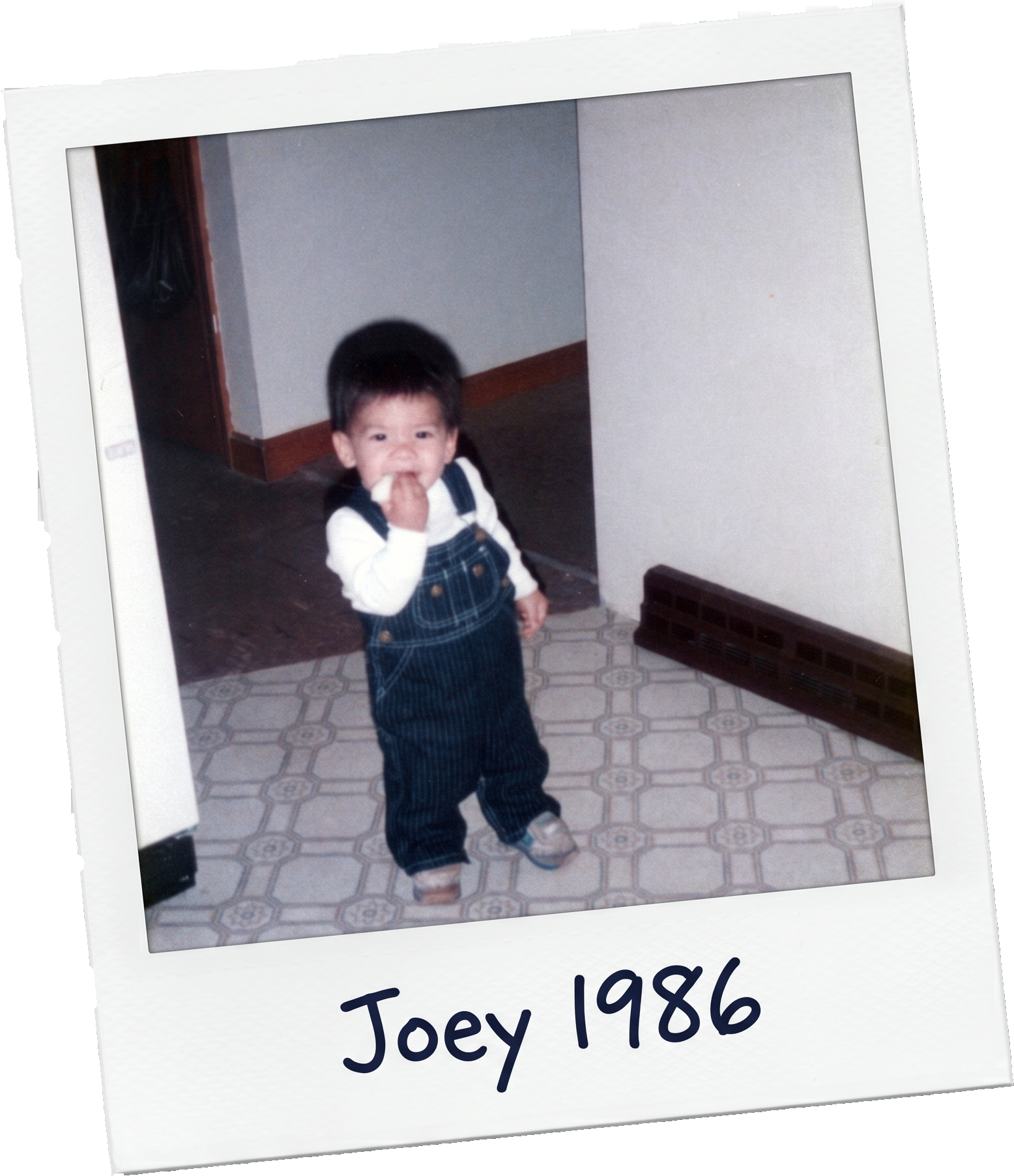 A young boy with dark hair wearing a white shirt and plaid overalls, standing on patterned floor tiles, with a wall heater behind him. The photo is labeled 'Joey 1986'.
