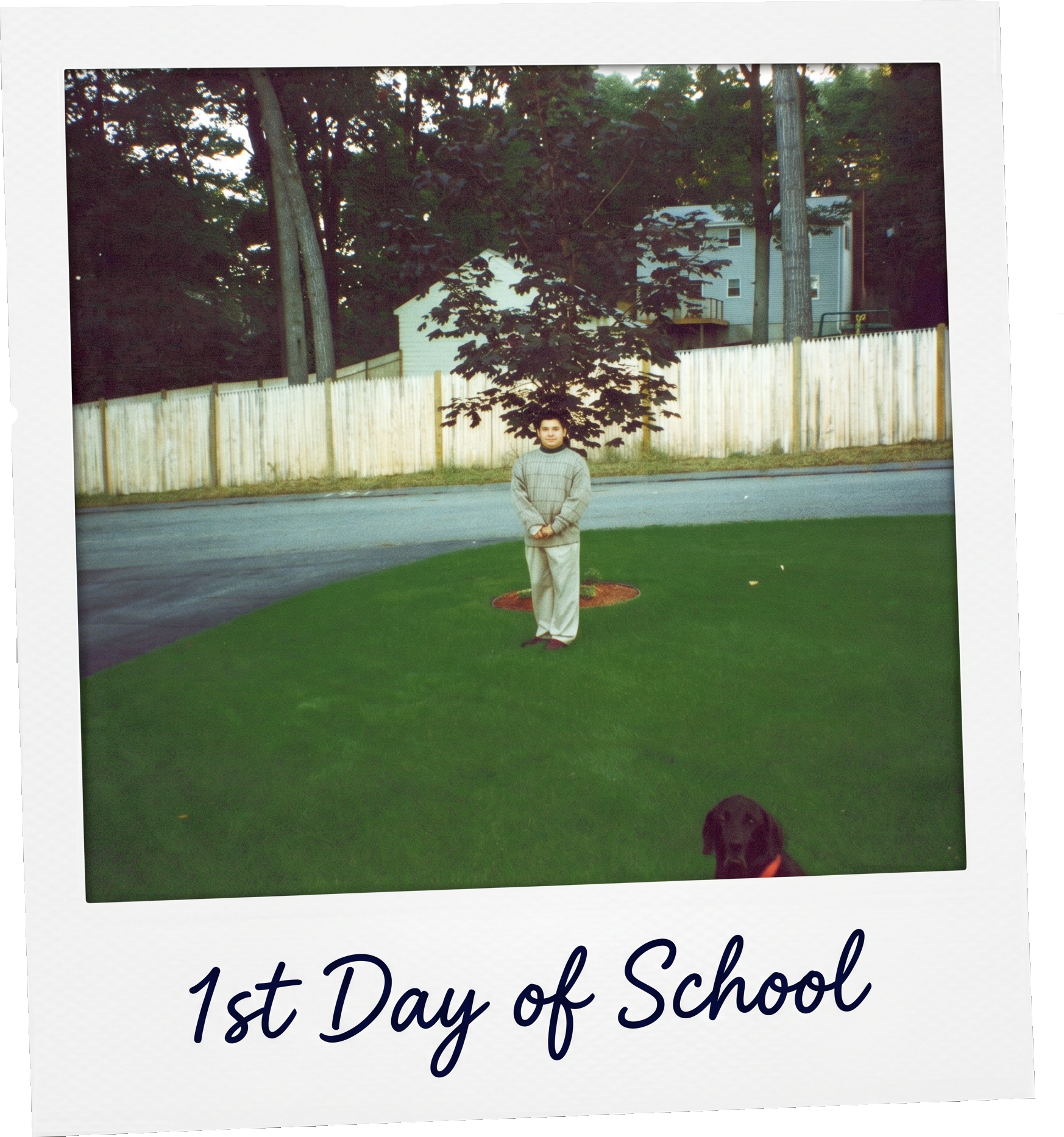 Photograph of a young boy standing on grass in front of a wooden fence, with trees and a house in the background. The boy is wearing a beige sweatshirt and light-colored pants. Part of a brown dog is visible at the bottom right corner. The image appears to be part of a scrapbook page, with the text "1st Day of School" at the bottom.