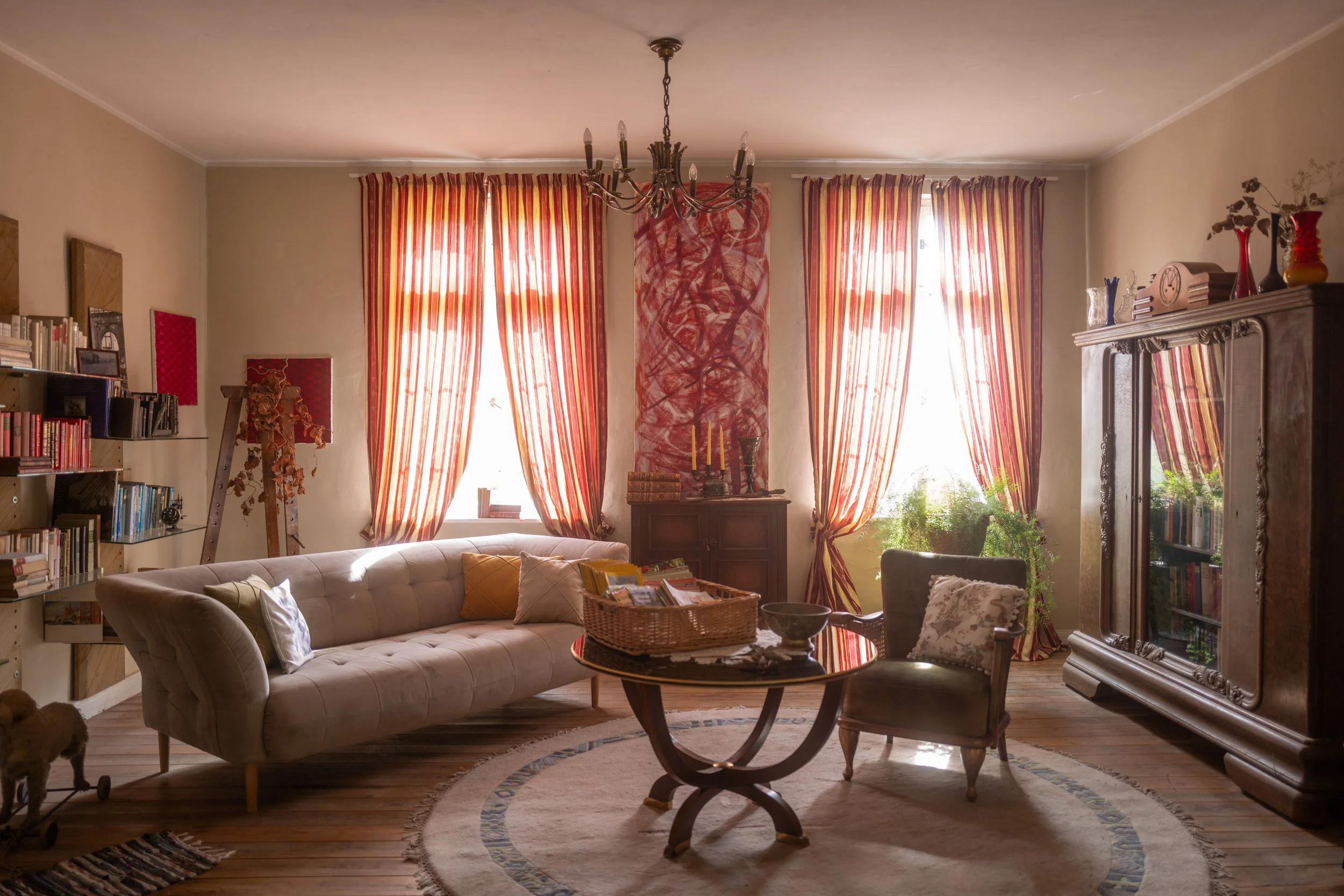Ein Wohnzimmer mit großen Fensterpanoramen, roten und gelb gestreiften Vorhängen, einer Stehlampe, einem Modernen Sessel, einem runden Teppich, Büchern und Deko-Objekten wie Vasen und Kerzen.