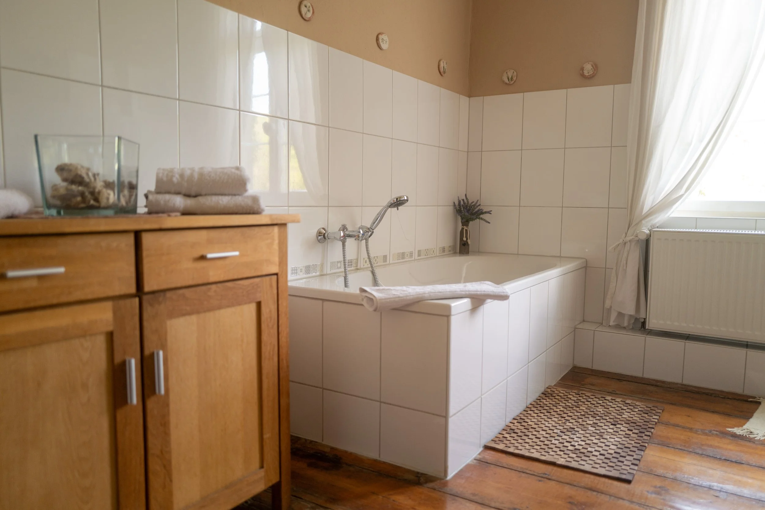 Ein Badezimmer mit einer Badewanne, weißen Fliesen an der Wand, einem Holzschrank, einem Handtuch und einem kleinen Strauß Lavendelpflanzen auf einer Flasche. Ein Fenster mit weißen Vorhängen lässt viel Licht ins Zimmer.