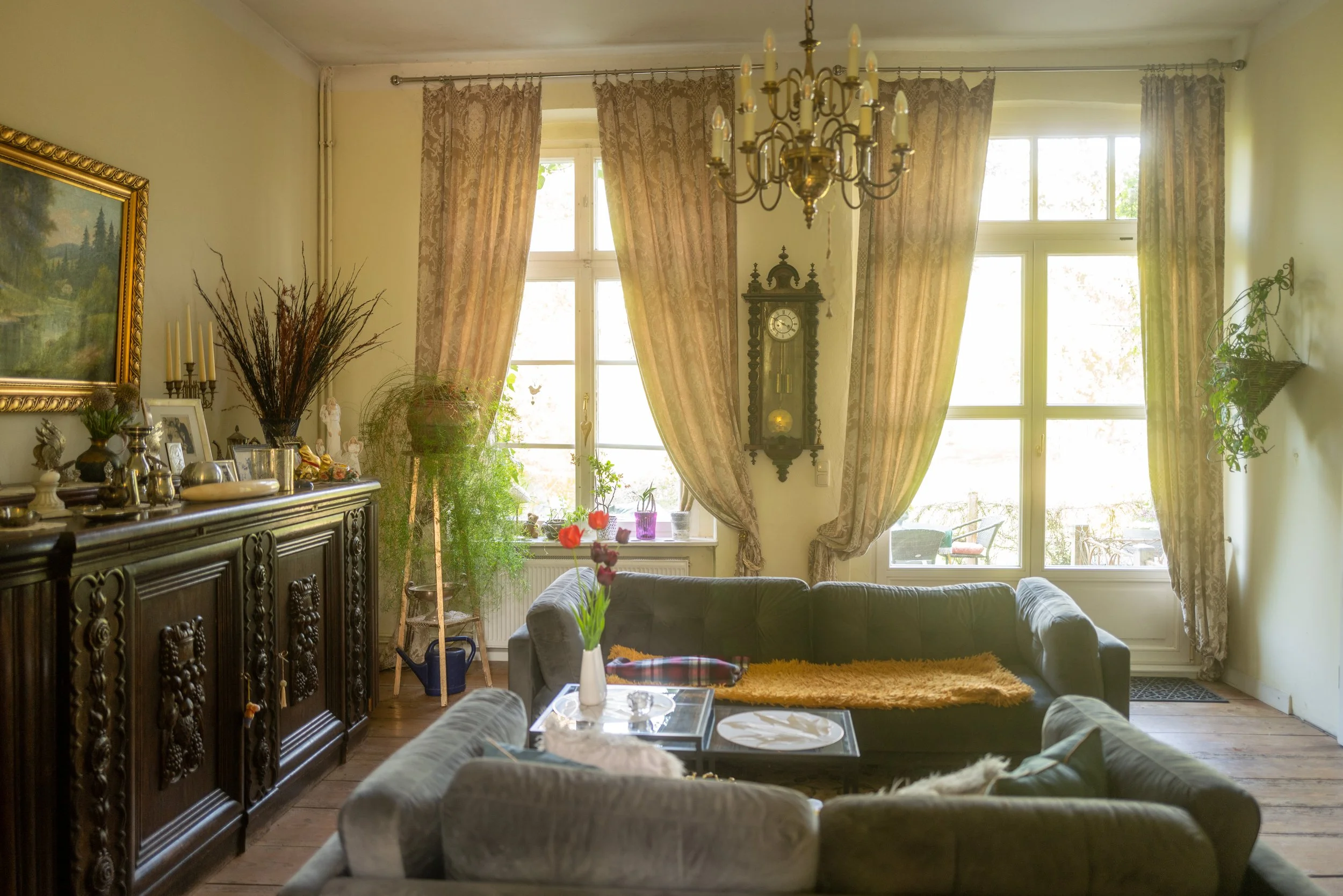 Wohnzimmer mit großen Fenstern, Vorhängen, einem alten Wanduhr, einem dunklen Sideboard mit Dekorationen, einer Couch, einem Glastisch mit Blumen und weiteren Pflanzen.