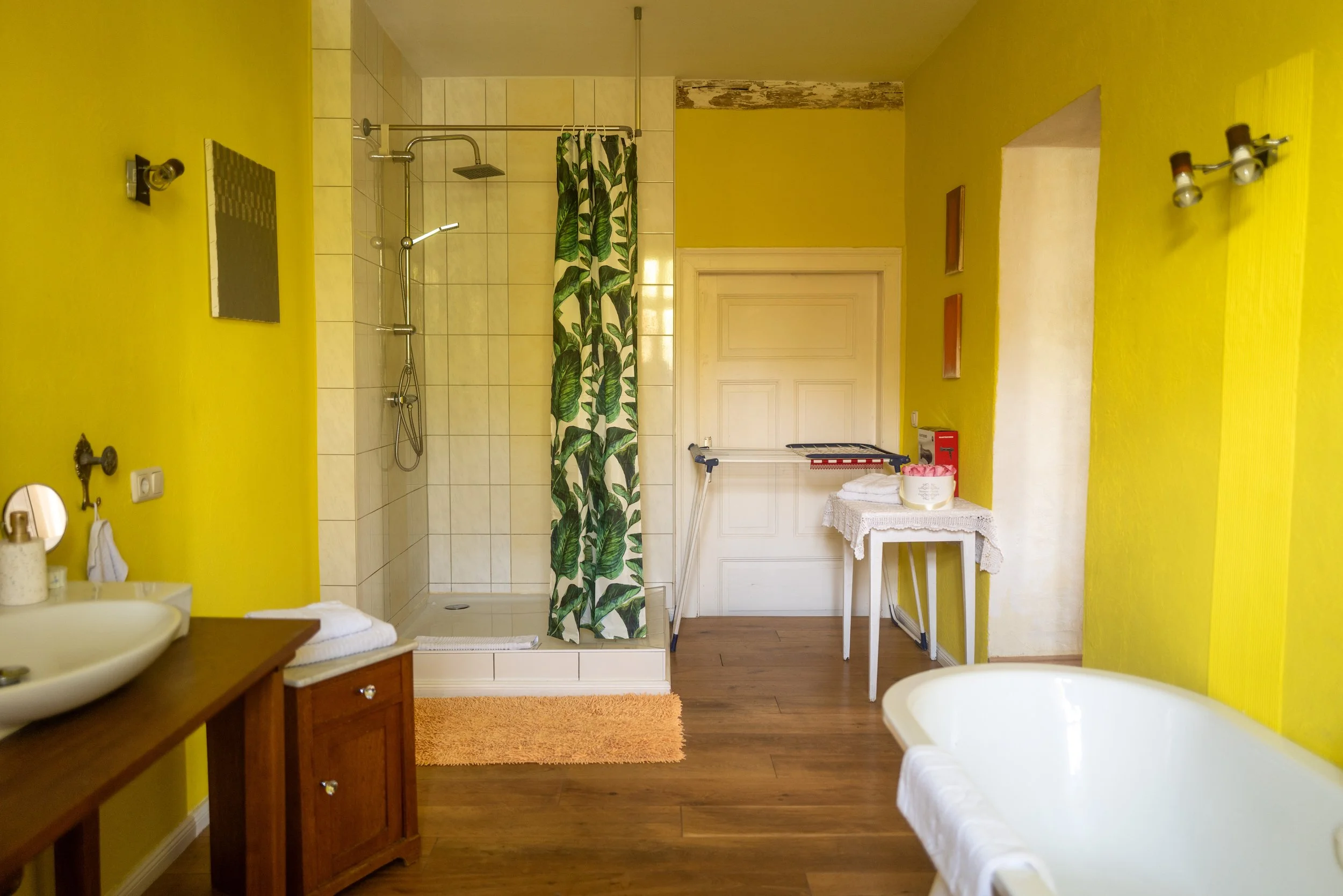 Innenansicht eines Badezimmers mit gelben Wänden, Dusche mit grün-blättrigem Duschvorhang, Holzfußboden, Waschbecken, Spiegel und Badewanne.