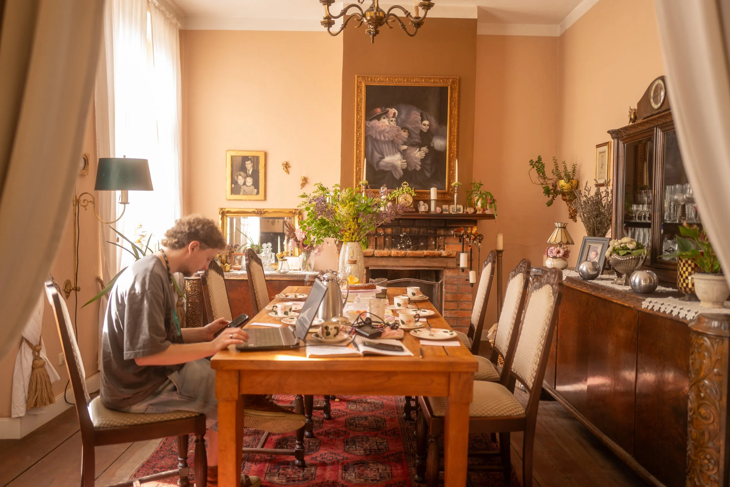 Ein Wohnzimmer mit einem großen Holztisch, auf dem Kaffeetassen, eine Lampe, Dekorationsgegenstände und ein Laptop stehen. Es gibt mehrere Stühle um den Tisch, eine Person sitzt und benutzt ein Smartphone. Im Hintergrund sind elegante Möbel, Bilder a