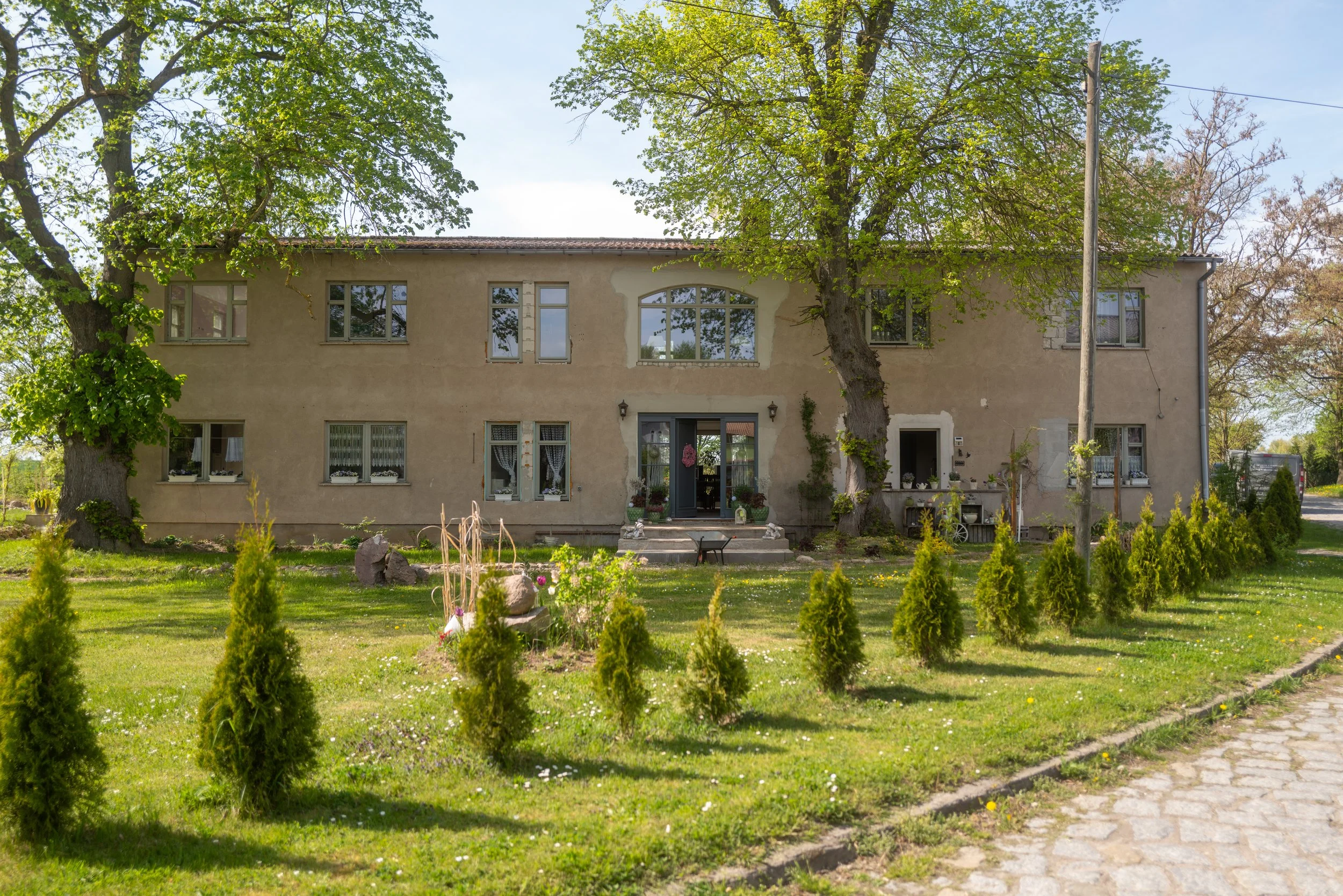 Ein zweistöckiges Haus mit einer beige Fassade, mehreren Fenstern und einem Eingang mit Deko im Garten. Zwei große Bäume spenden Schatten, im Vordergrund ist eine gepflegte Rasenfläche mit kleinen Sträuchern und einem Blumenbeet.