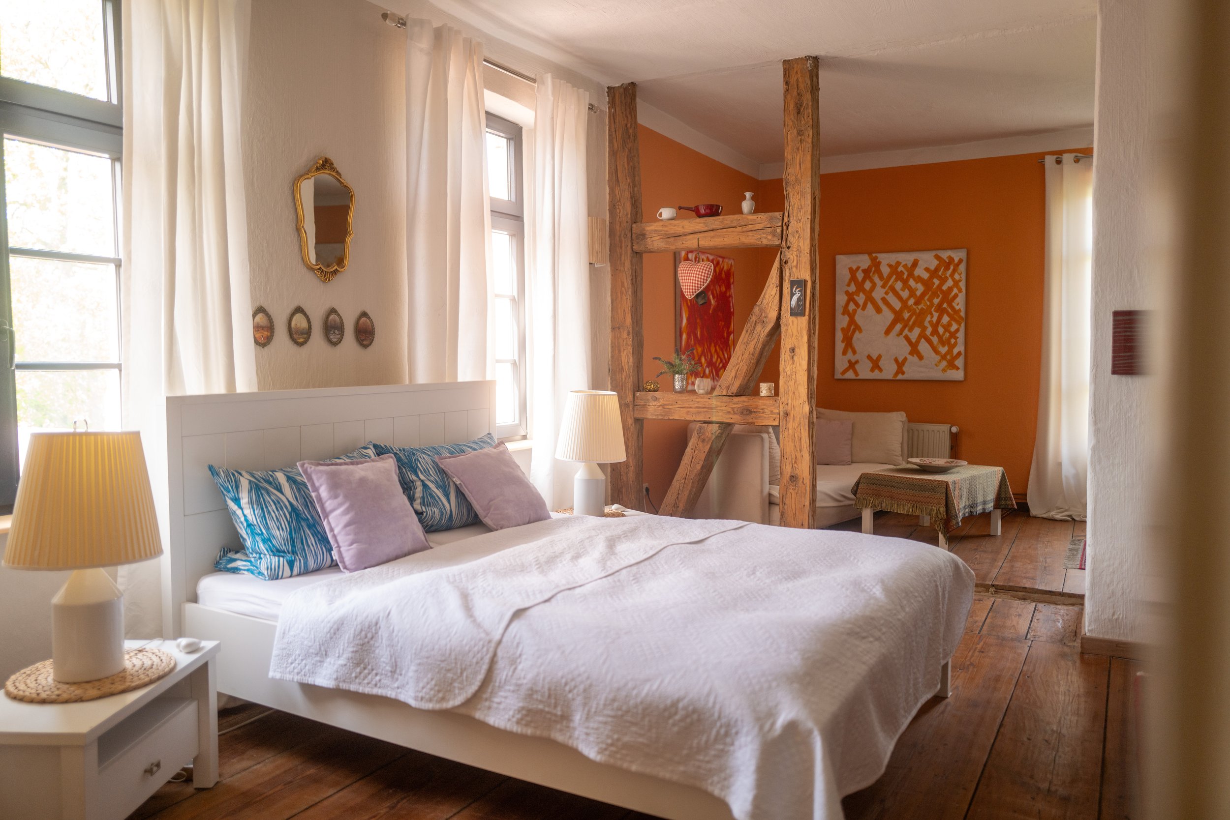 Ein Schlafzimmer mit einem weißen Bett, mehreren Kissen, Nachttischlampe, großen Fenstern mit weißen Vorhängen, einer Holztrennwand mit dekorativen Gegenständen und bunten Wandbildern, einem orangefarbenen Wandhintergrund und Holzboden.