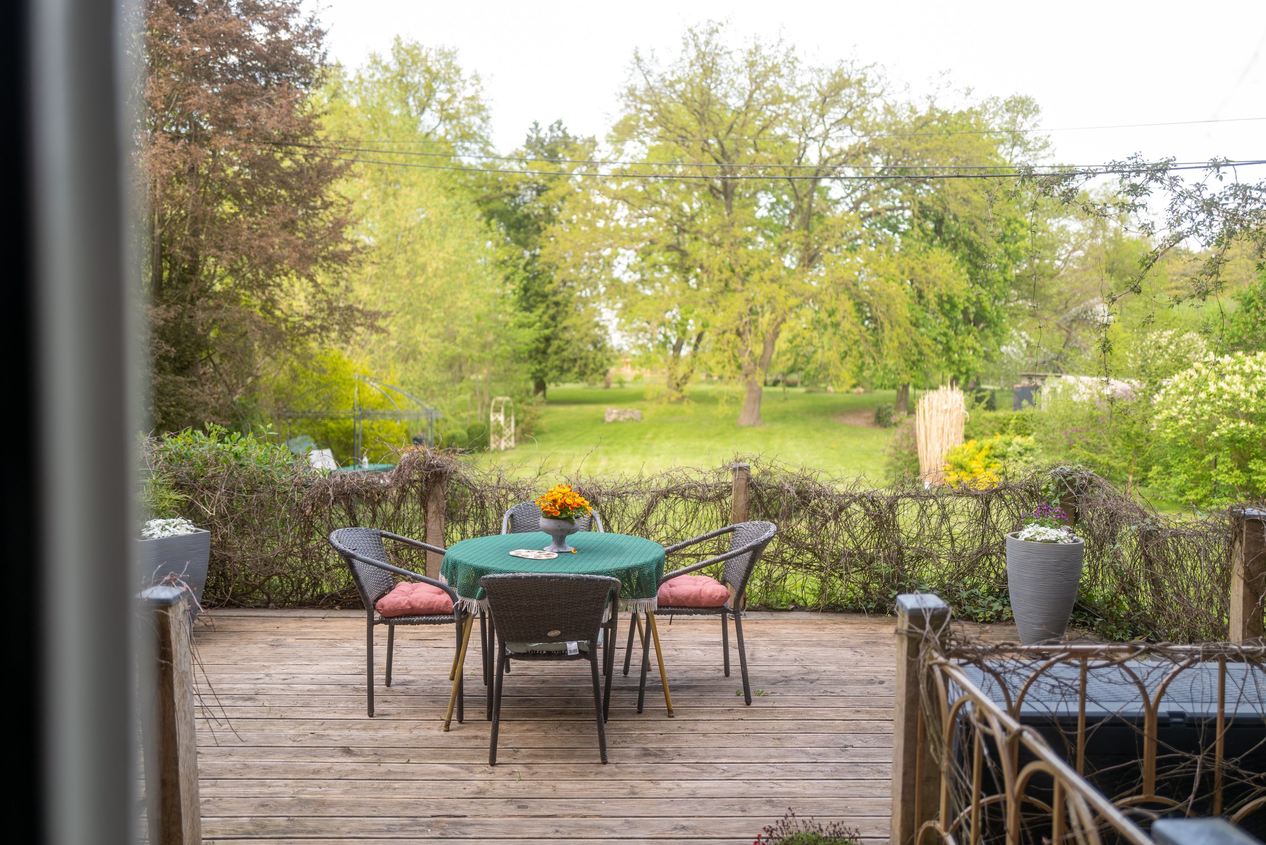Blick auf eine Garten-Terrasse mit runder Tafel, sechs Stühlen und Topfpflanzen, mit Blick auf einen grünen Garten mit Bäumen und Gras. Kommt durch ein geöffnetes Fenster.