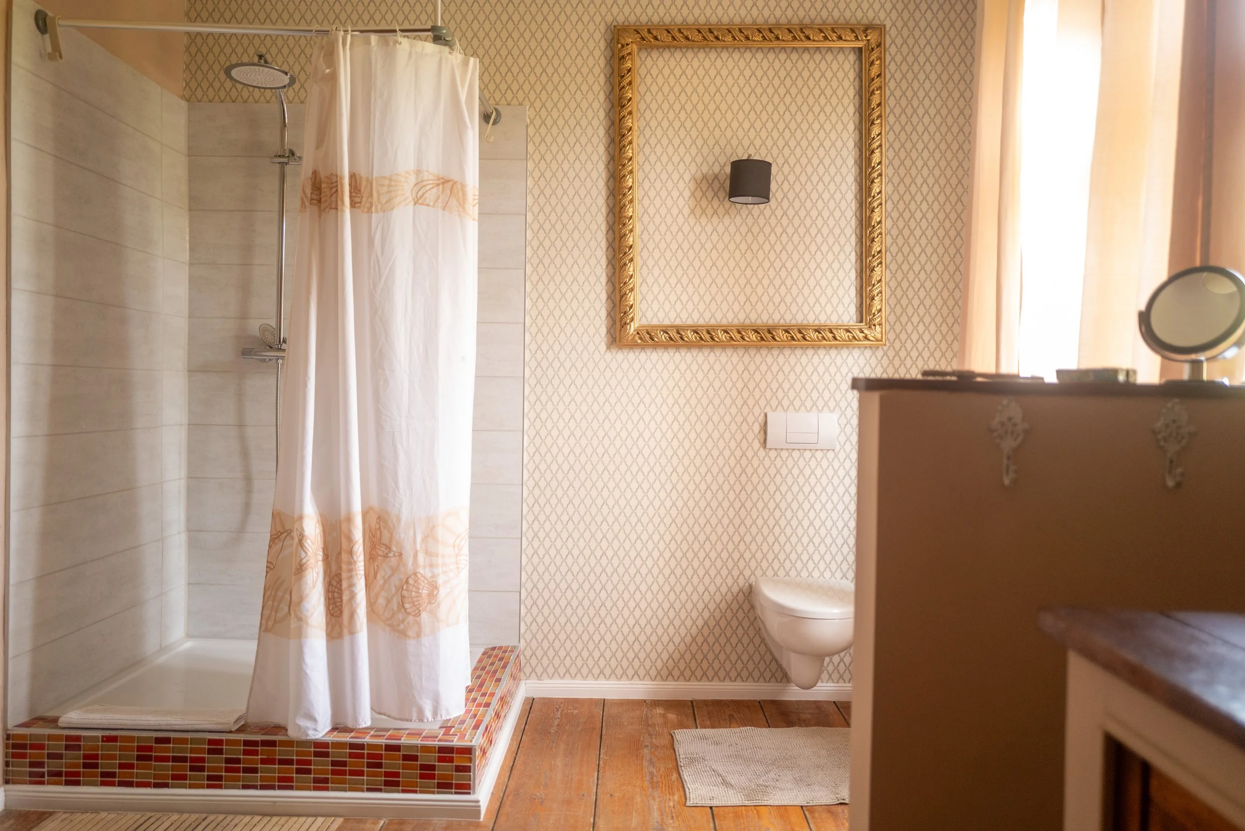 Innenansicht eines Badezimmers mit Dusche, Toilette, Spiegel, schwarzem Lampenschirm und einem Fenster mit halbtransparenten Vorhängen.