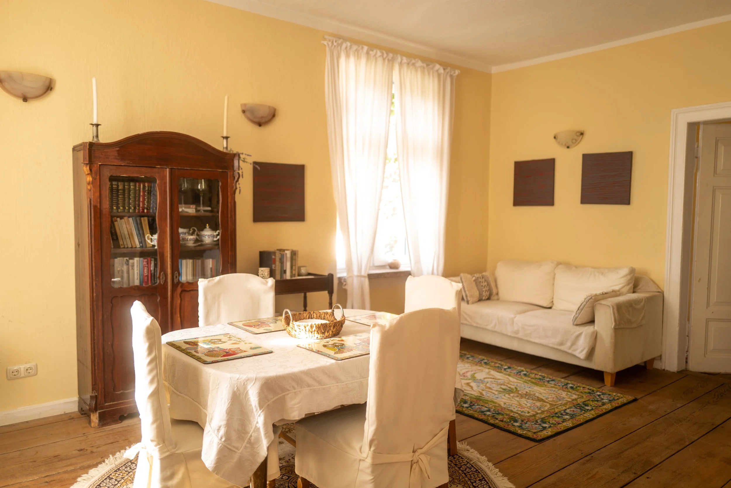 Ein Wohnzimmer mit gelben Wänden, weißen Vorhängen, einer Couch und einem Essbereich, einer alten Holzvitrine mit Büchern und Geschirr, einem gewebten Teppich und Wanddekorationen.