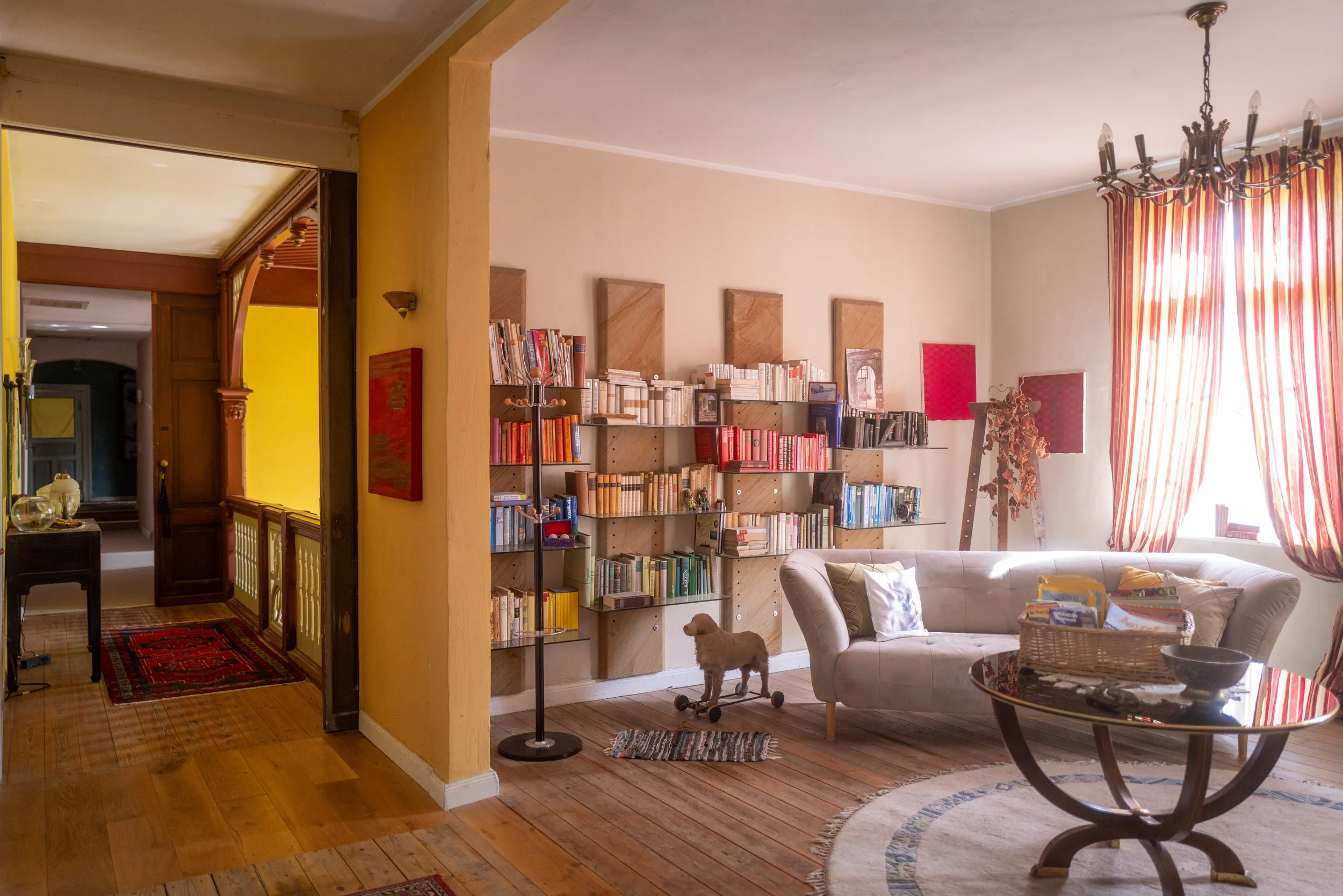 Ein Wohnzimmer mit einem Bücherregal, einem weißen Sofa, einem Couchtisch, einer Gardine mit roten Streifen und einem Kronleuchter.