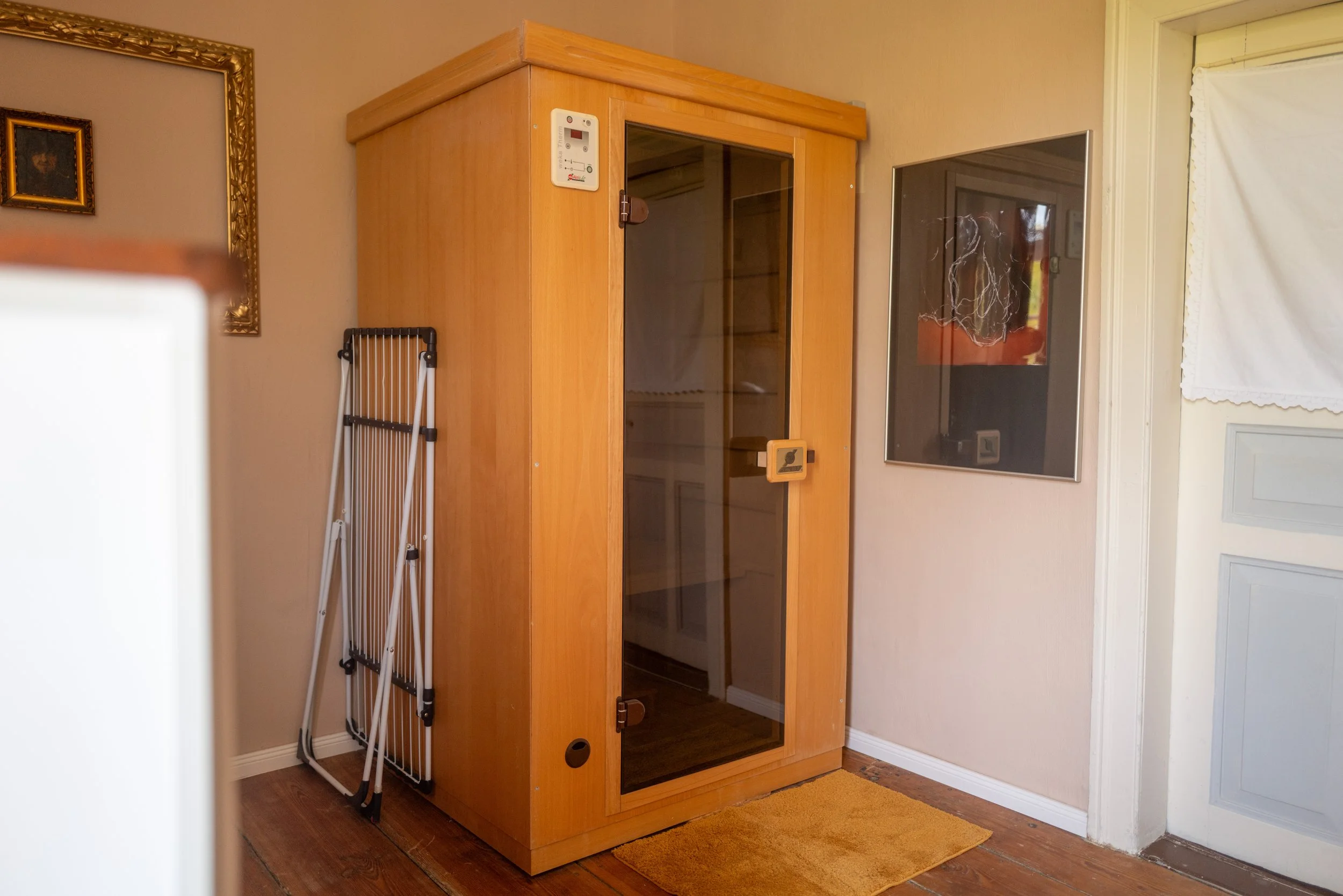 Ein holzfarbener Infrarot-Kabine mit Glasfront, neben einem Wandbild, einem zusammenklappbaren Wäscheständer und einem Teppich vor einer weißen Wand.