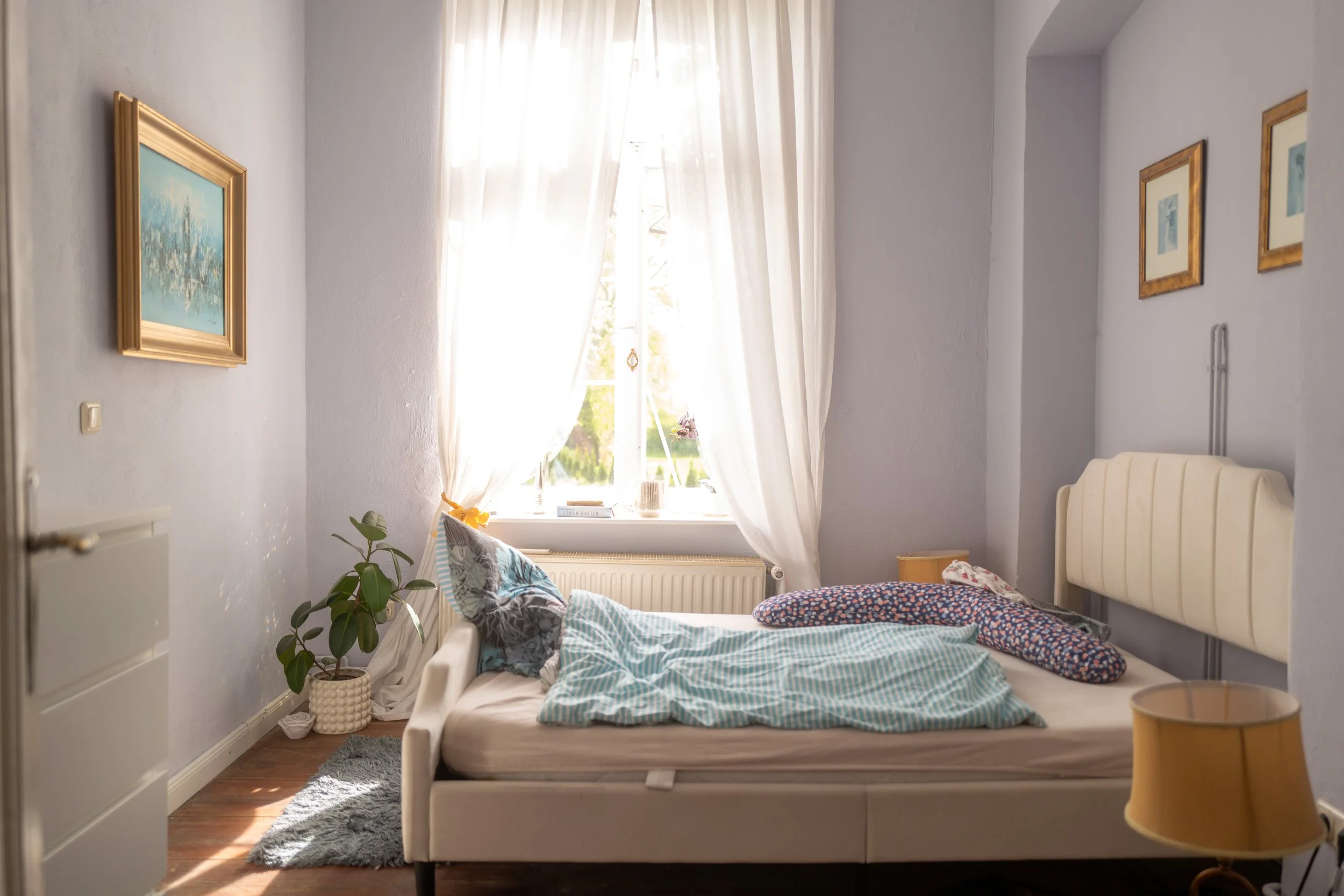 Ein unordentliches Schlafzimmer mit einem offenen Bett, das Bettdecke und Kissen hat. Es gibt einen großen Fenster mit weißen Vorhängen, durch die Tageslicht scheint. An den Wänden hängen Bilder, und es steht eine grüne Pflanze in einem Korb neben dem Bett.