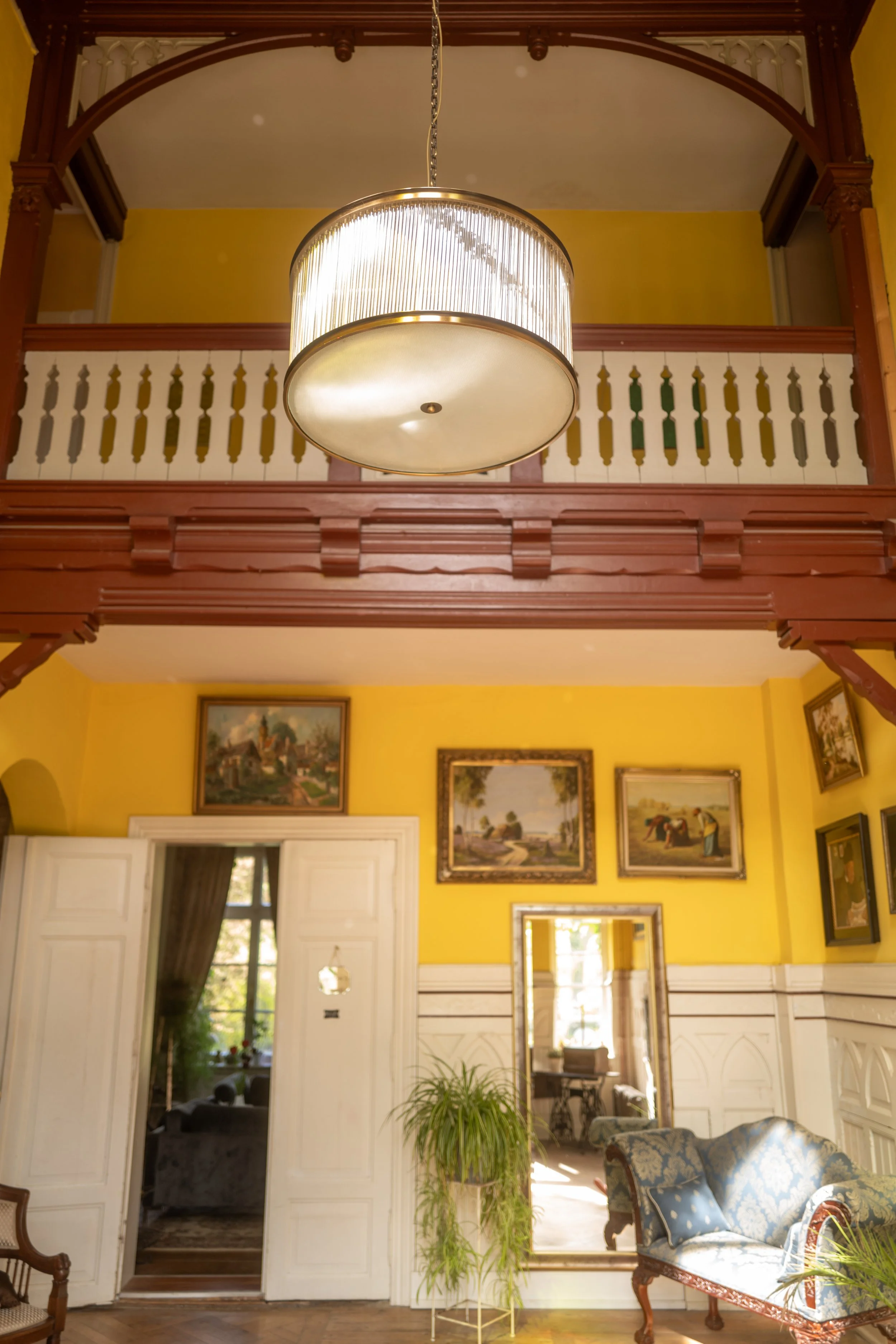 Interior eines historischen Raumes mit gelben Wänden, einem Kronleuchter, Gemälden an der Wand, einem offenen weißen Tür, einem blauen Sitzmöbel und Bildern im Hintergrund.