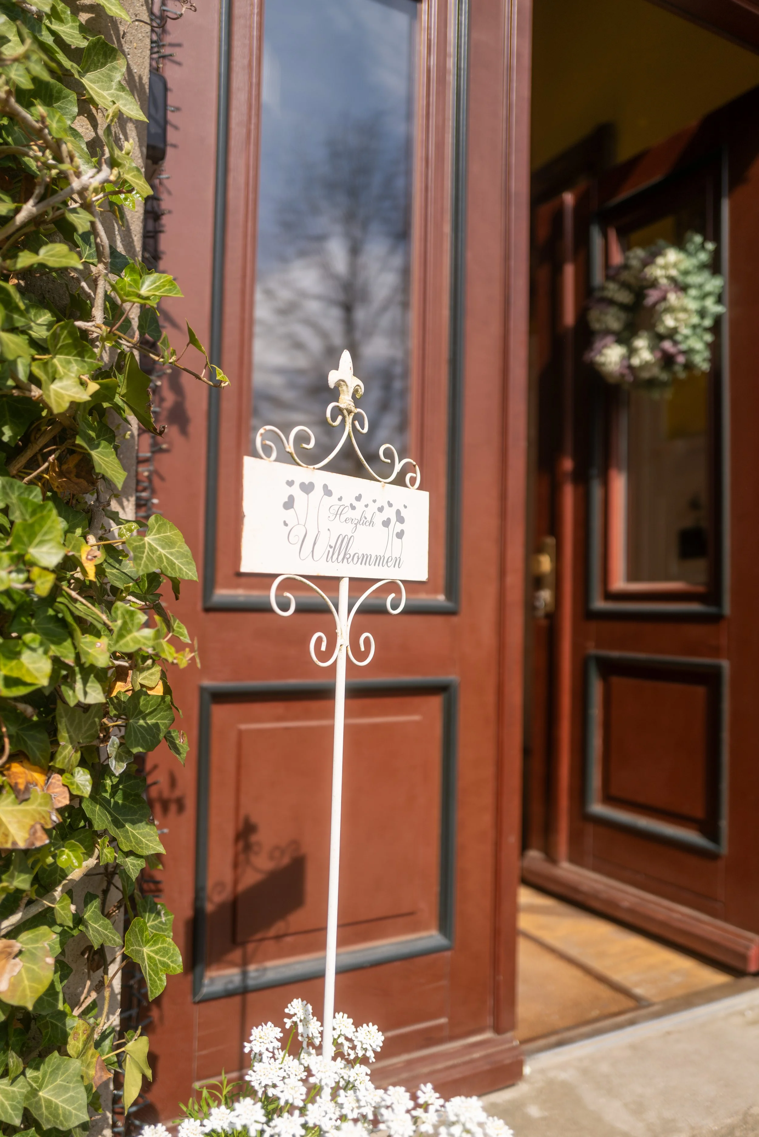 Der Eingang eines Hauses mit einer roten Holztür, umgeben von grünen Efeu-Pflanzen. Im Vordergrund befindet sich ein weißer Willkommensschild auf einem Pfahl, mit weißen Blumen am unteren Rand. Das Haus hat eine offene Tür, durch die ein Kranz aus we