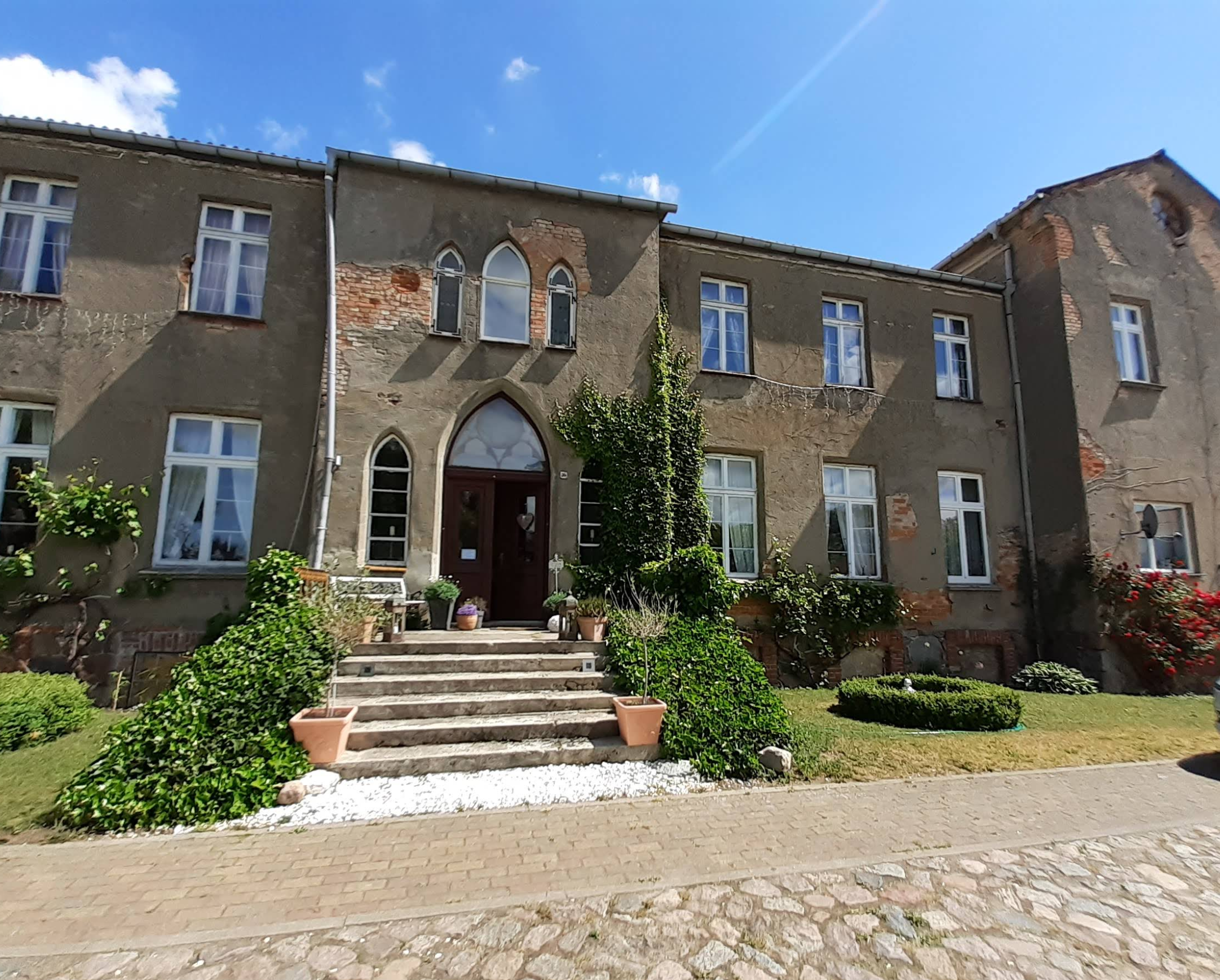 Eine alte Gebäude mit mehreren Fenstern und einer Treppe, die zum Eingang führt, im Garten mit Pflanzen, unter blauem Himmel mit wenigen Wolken.