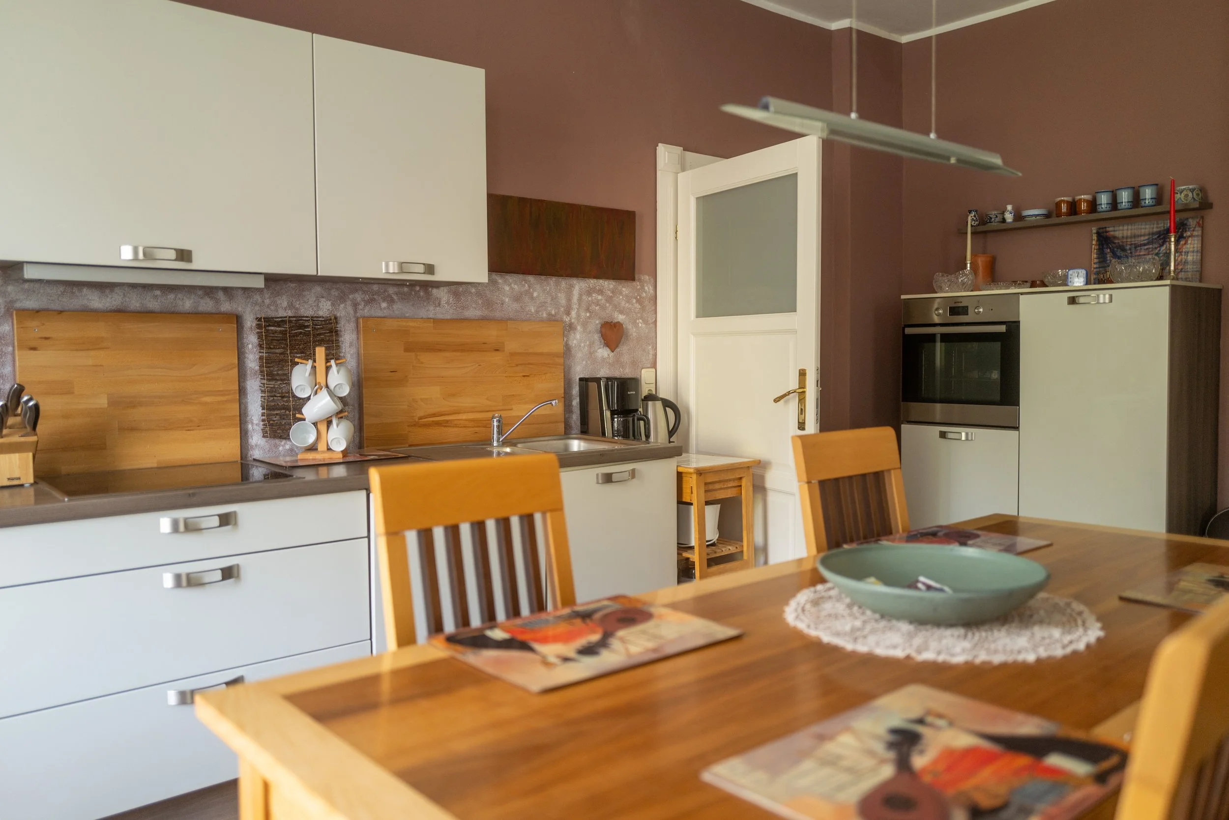 Ein Blick in eine gemütliche Küche mit weißen Schränken, Holzregalen, Kühlschrank, Herd und Esstisch mit Stühlen, sowie verschiedenen Küchengeräten und Dekorationen.