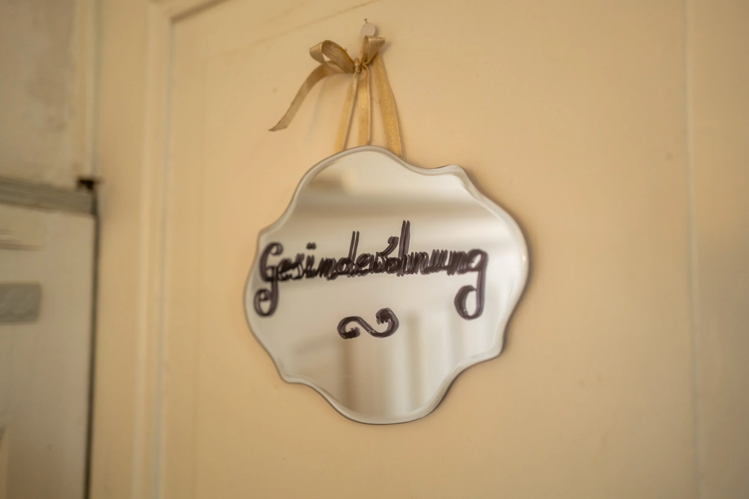 Ein ovaler Wandspiegel mit einer goldenen Schleife am oberen Rand, der an einer cremefarbenen Wand hängt. Darunter ist eine dekorative Tafel mit handgeschriebener Aufschrift, die teilweise sichtbar ist.