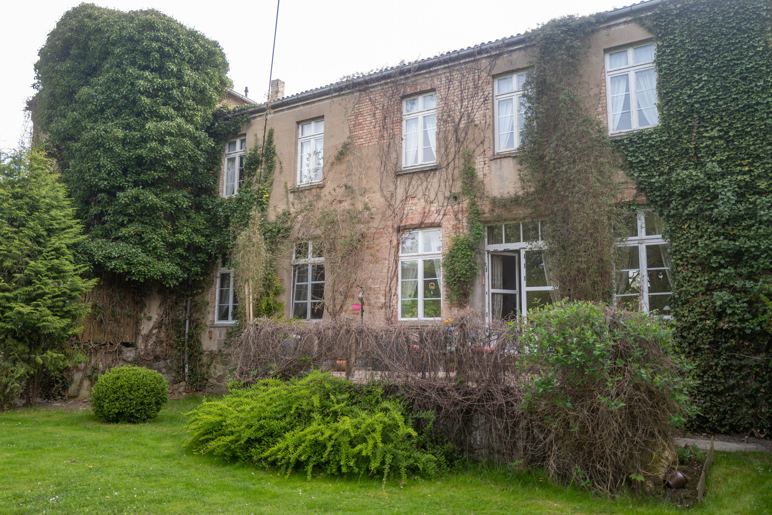 Ein Haus mit hohen Fenstern, das teilweise von Efeu und Kletterpflanzen überwachsen ist, umgeben von einem grünen Garten mit Büschen und Rasen.