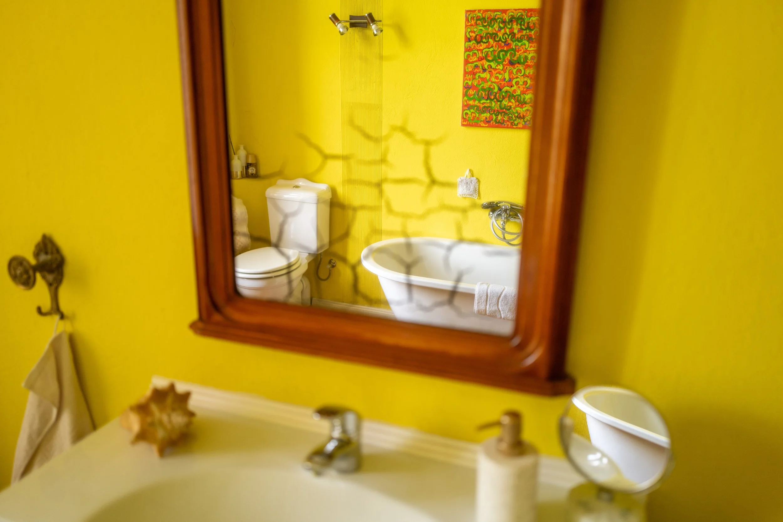 Ein Badezimmer mit gelber Wandfarbe, ein Spiegel, eine Toilette, ein Waschbecken, ein Handtuch und ein Kunstwerk an der Wand.