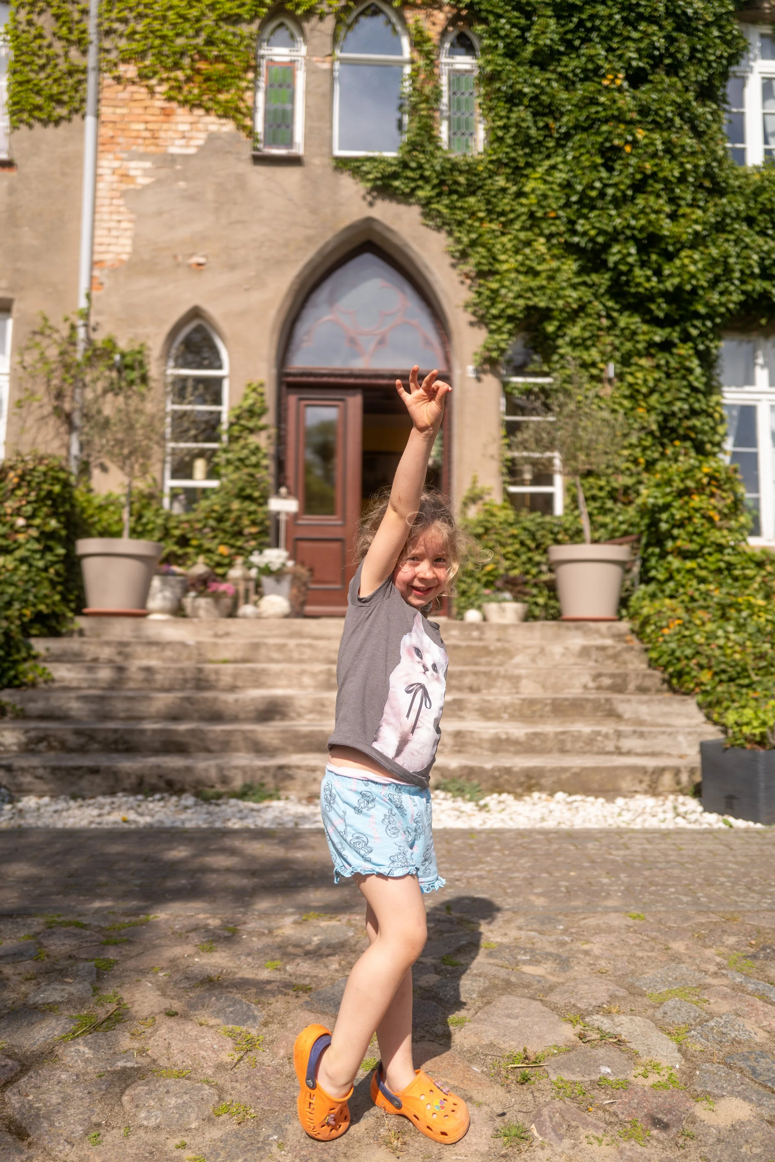 Ein Mädchen steht vor einem alten Gebäude mit Steintreppe, grünen Pflanzen an den Wänden und hohen Fenstern, hebt den Arm und zeigt die Handzeichen, trägt eine Graue T-Shirt mit Katzenmotiv, Shorts mit Meerestieren, orange Crocs.
