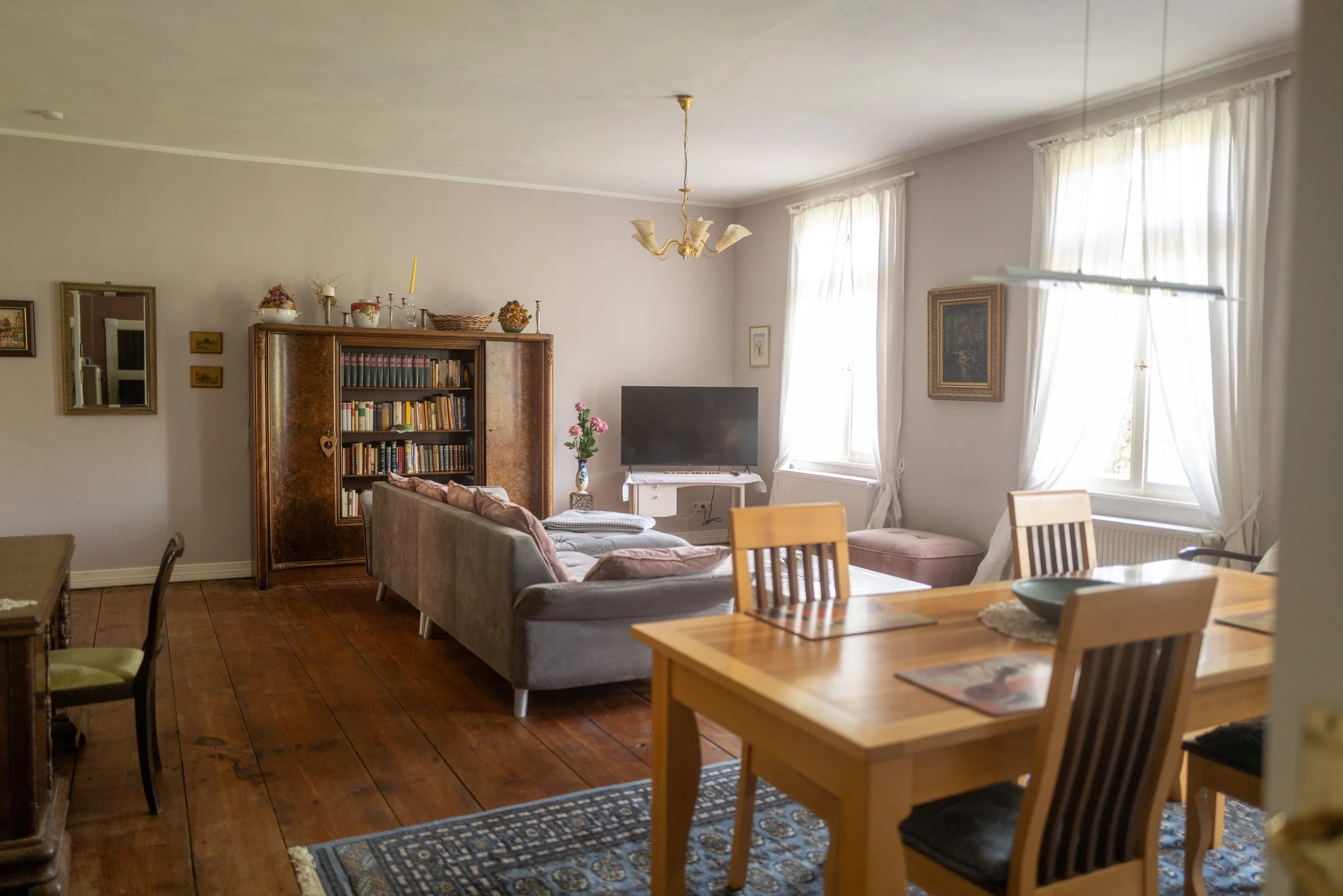Ein Wohnzimmer mit einem großen Sofa, einem Fernseher, einem Bücherregal, mehreren Fenstern mit weißen Vorhängen, einem Kronleuchter an der Decke und verschiedenen Dekorationsgegenständen auf dem Regal.