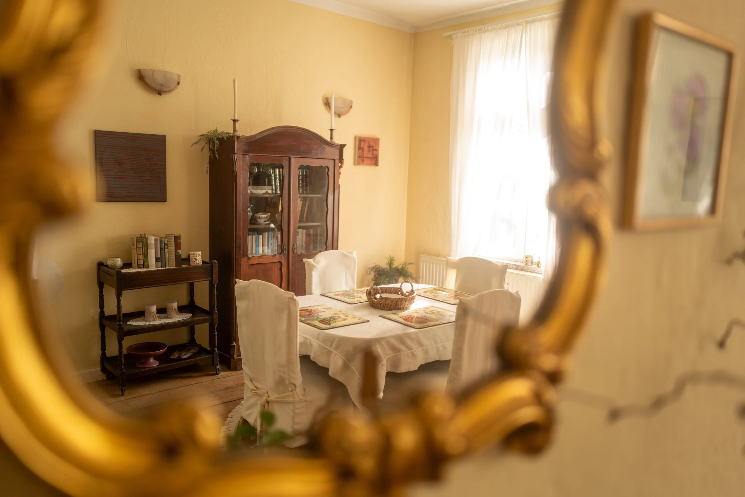 Einblick in ein gemütliches Esszimmer, sichtbar durch eine goldene, verzierte Spiegelrahmenöffnung. Der Raum hat gelbe Wände, weiße Vorhänge, einen Holzboden und einen Esstisch mit weißen Stühlen. Im Hintergrund ein dunkler Bücherregal-Kabinett, Wand
