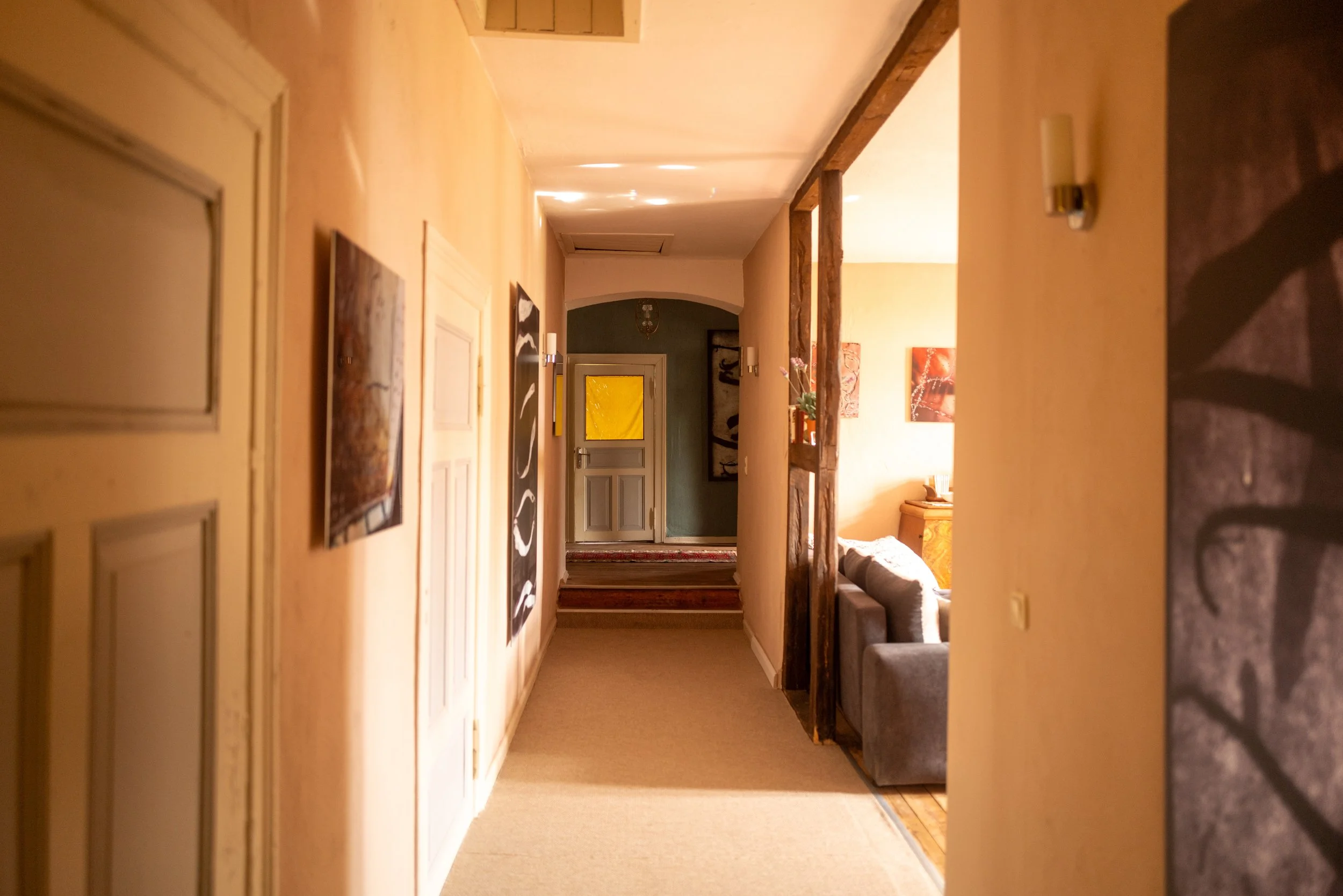 Innenaufnahme eines Flurs mit mehreren Bildern an der Wand, einer Holzkonstruktion, einem Sofa im Hintergrund und dekorativen Beleuchtungen.