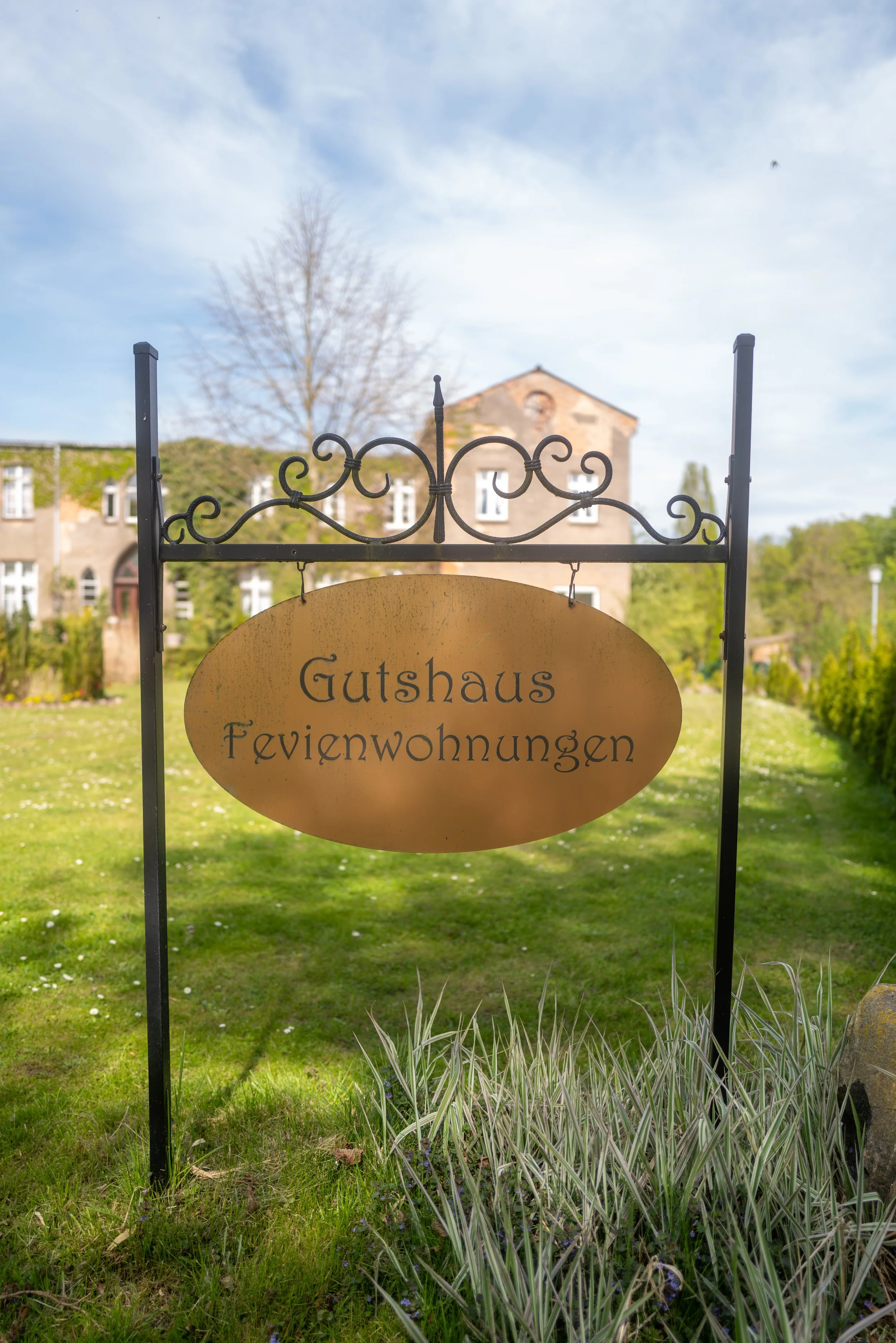 Hinweisschild mit der Aufschrift 'Gutshaus Ferienwohnungen' vor einem grünen Garten mit Büschen und Bäumen, im Hintergrund ein historisches Gebäude und blauer Himmel mit Wolken.