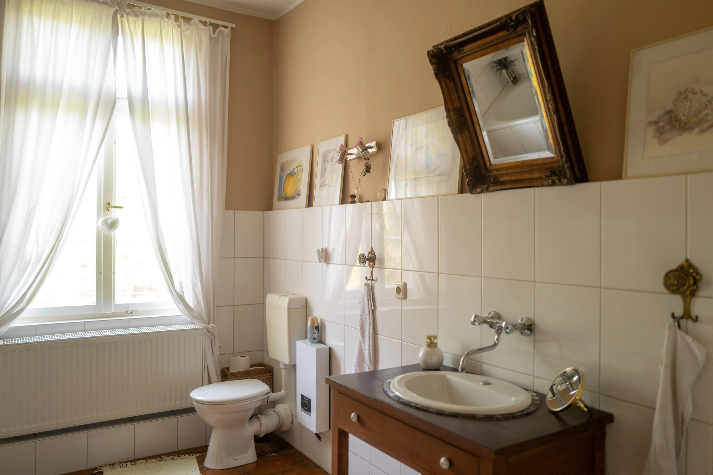 Ein Badezimmer mit einem Fenster mit weißen Vorhängen, einem weißen WC, einem Waschbecken auf einem Holzunterschrank, einem großen Wandspiegel mit einer alten goldfarbenen Rahmen, drei Bilder an der Wand, einem Kasten mit Toilettenpapier, einer kleinen Waschmaschine, einem Herz- dekorativen Gegenstand am Fenstergriff und einer Heizkörper darunter.