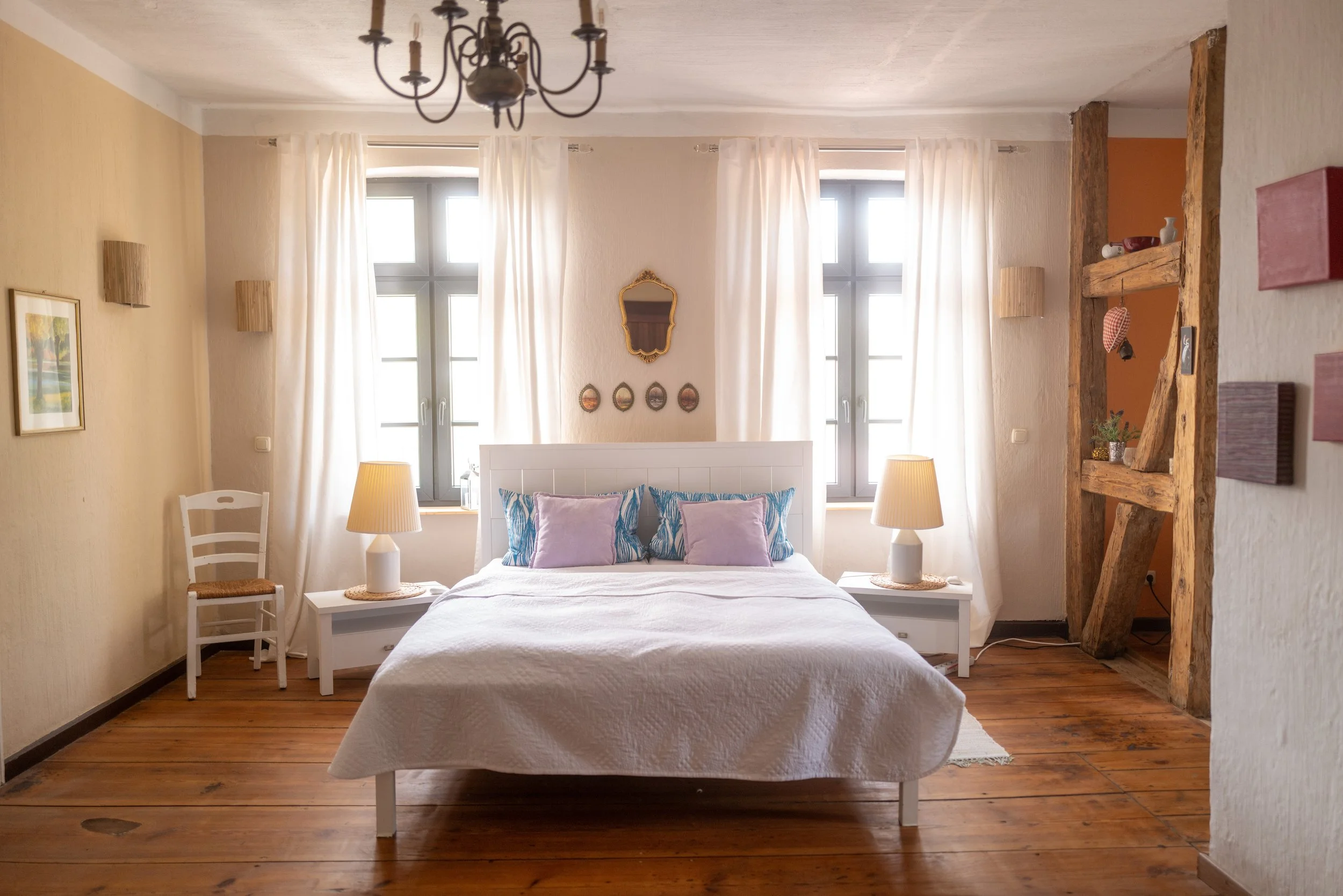 Ein gemütliches Schlafzimmer mit weißem Bett, blauen und lilafarbenen Kissen, zwei Nachttischen mit Lampen, großen Fenstern mit weißen Vorhängen, an der Wand ein goldgerahmter Spiegel und dekorative Wandteile, eine antike Wanduhr, Blick auf Holzfußbo