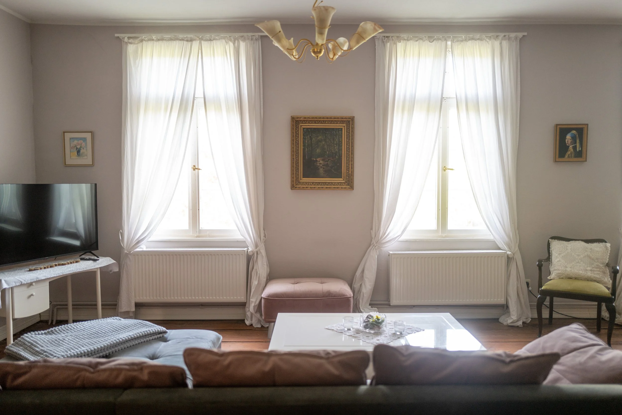 Wohnzimmer mit weißen Vorhängen, antiker Lampe, Bilder an der Wand, Fernseher, Sofa mit Kissen, Couchtisch mit Dekoration, Stuhl, gepolsterte Bank, Heizung unter den Fenstern.